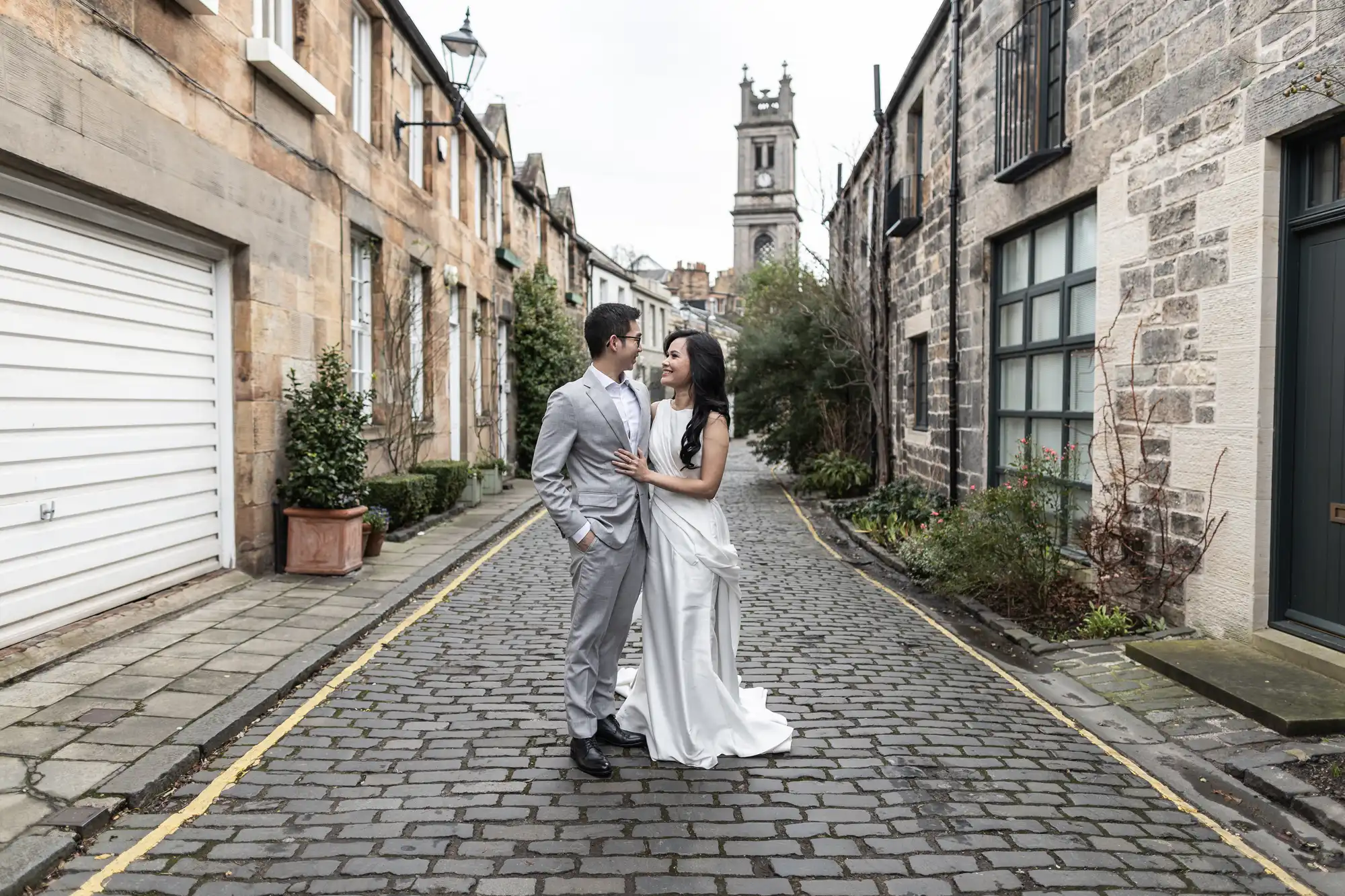 A couple dressed in formal wedding attire stand close together on a cobblestone street with stone buildings and a clock tower in the background.