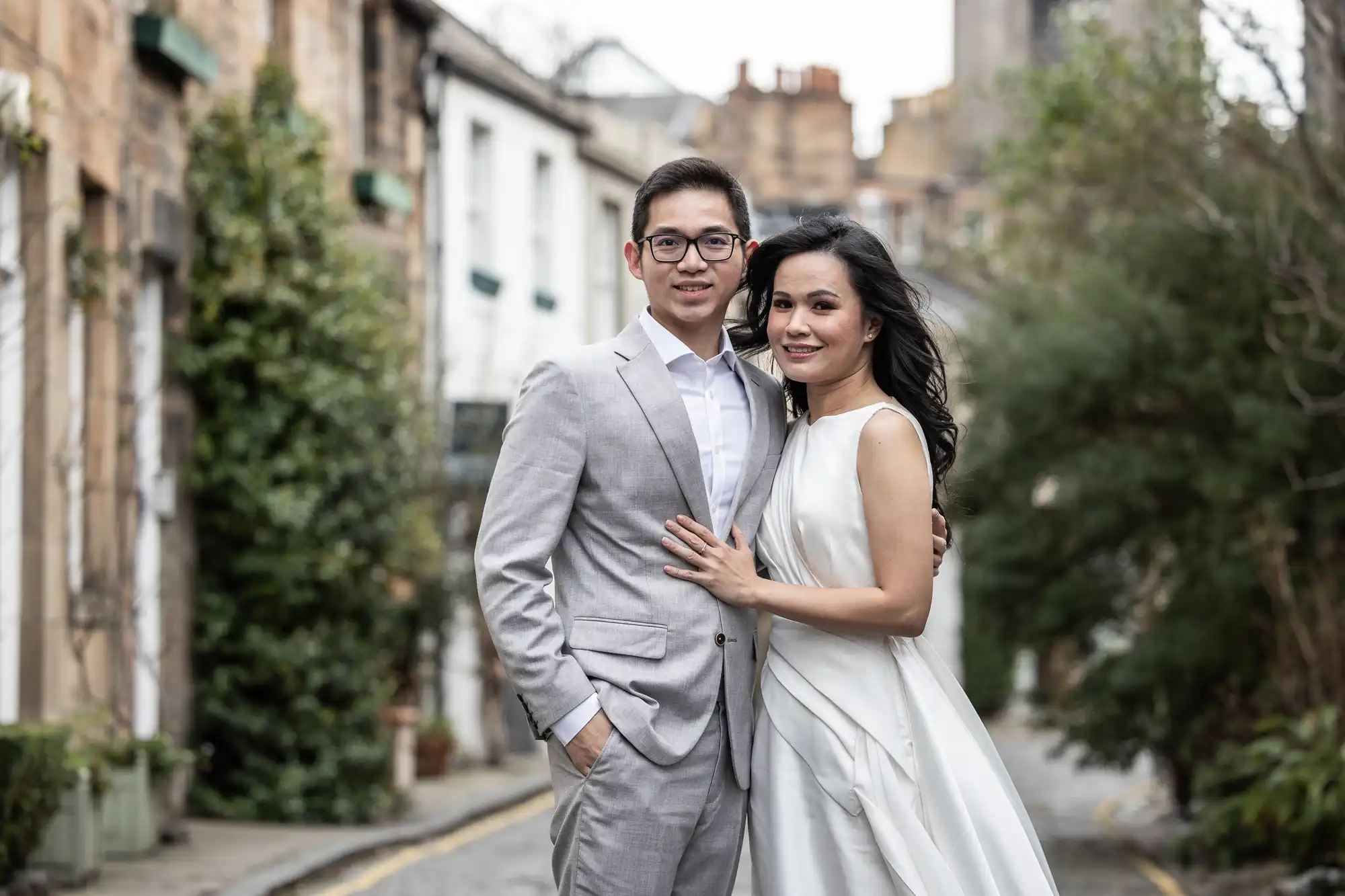 A man in a light grey suit and a woman in a white dress stand closely together, smiling, on a narrow street lined with buildings and trees.