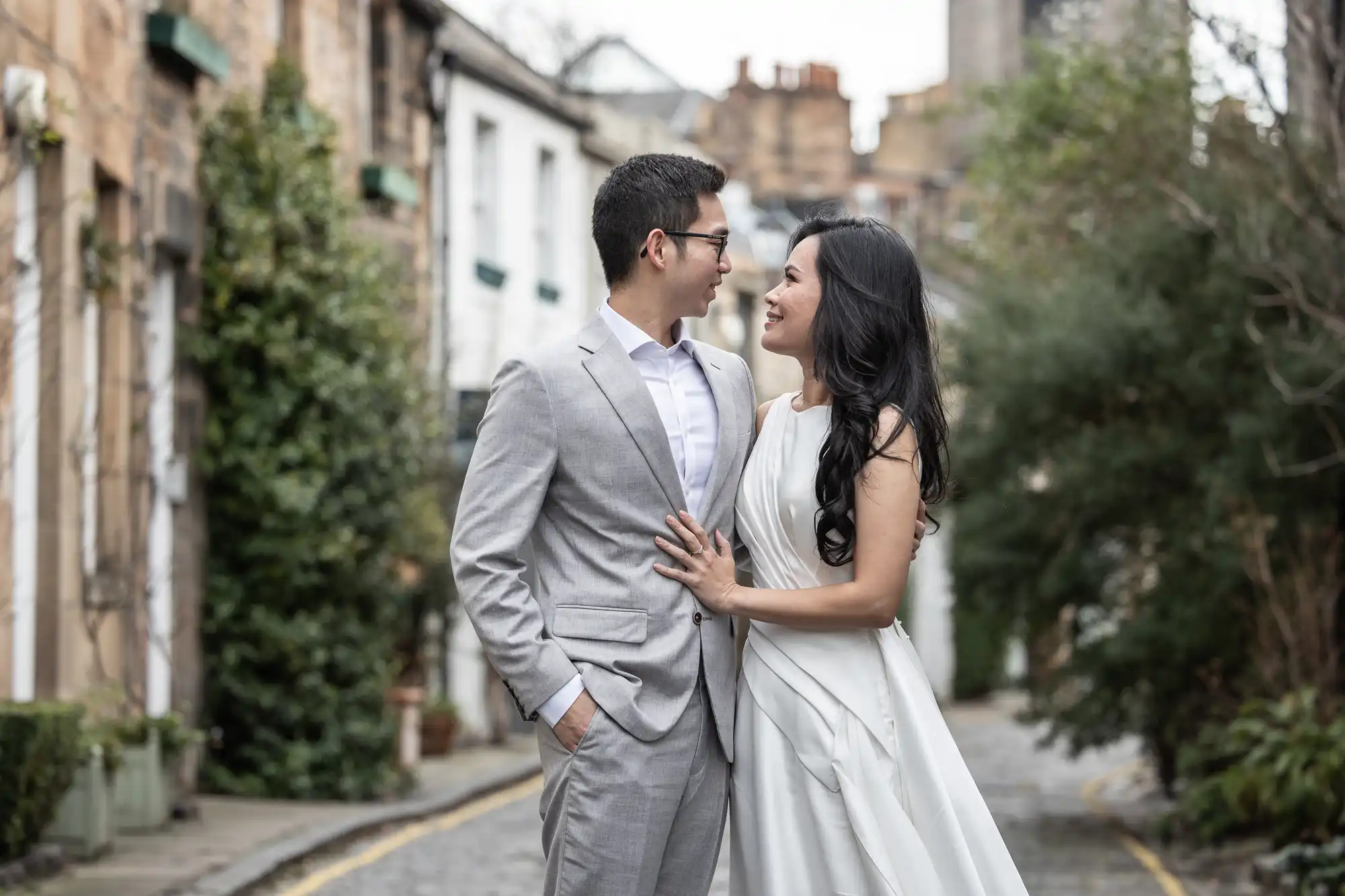 A couple stands and smiles at each other in an outdoor setting, both dressed formally; the man in a grey suit and the woman in a white dress. The background shows greenery and buildings.