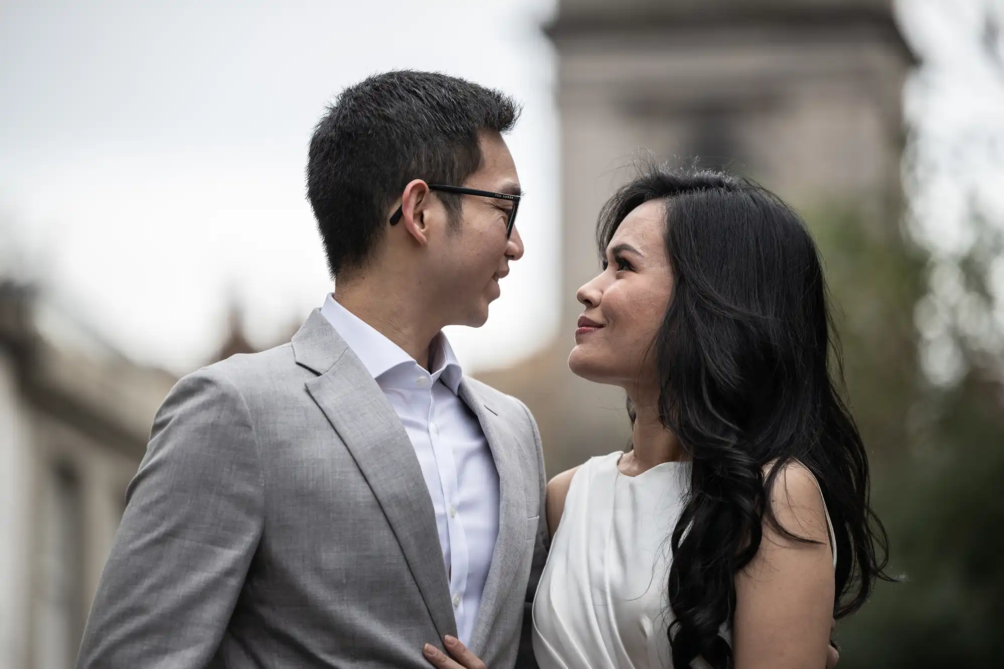 A man in a gray suit and glasses and a woman in a white dress are standing closely, looking at each other with smiles, outdoors.