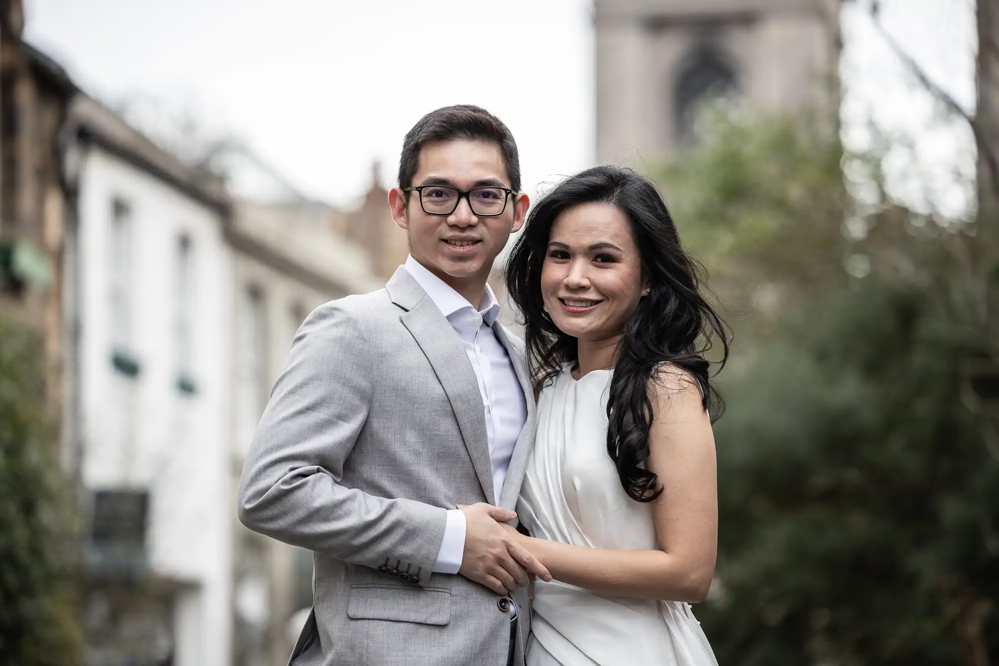 A man in a light gray suit and a woman in a white dress stand closely together and smile outdoors in front of a blurred architectural background.