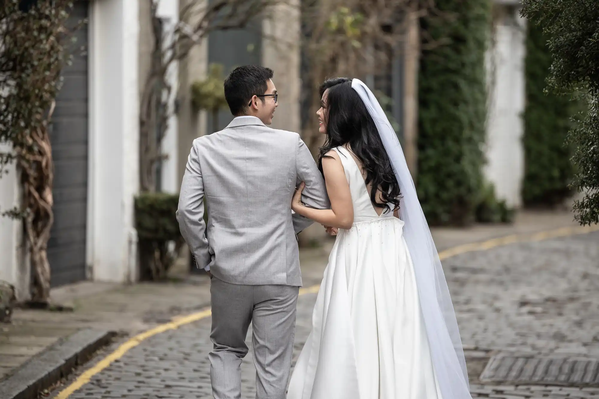 A bride and groom walk arm in arm down a cobblestone path. She is in a white dress and veil, and he is in a light gray suit. They are looking at each other and smiling.