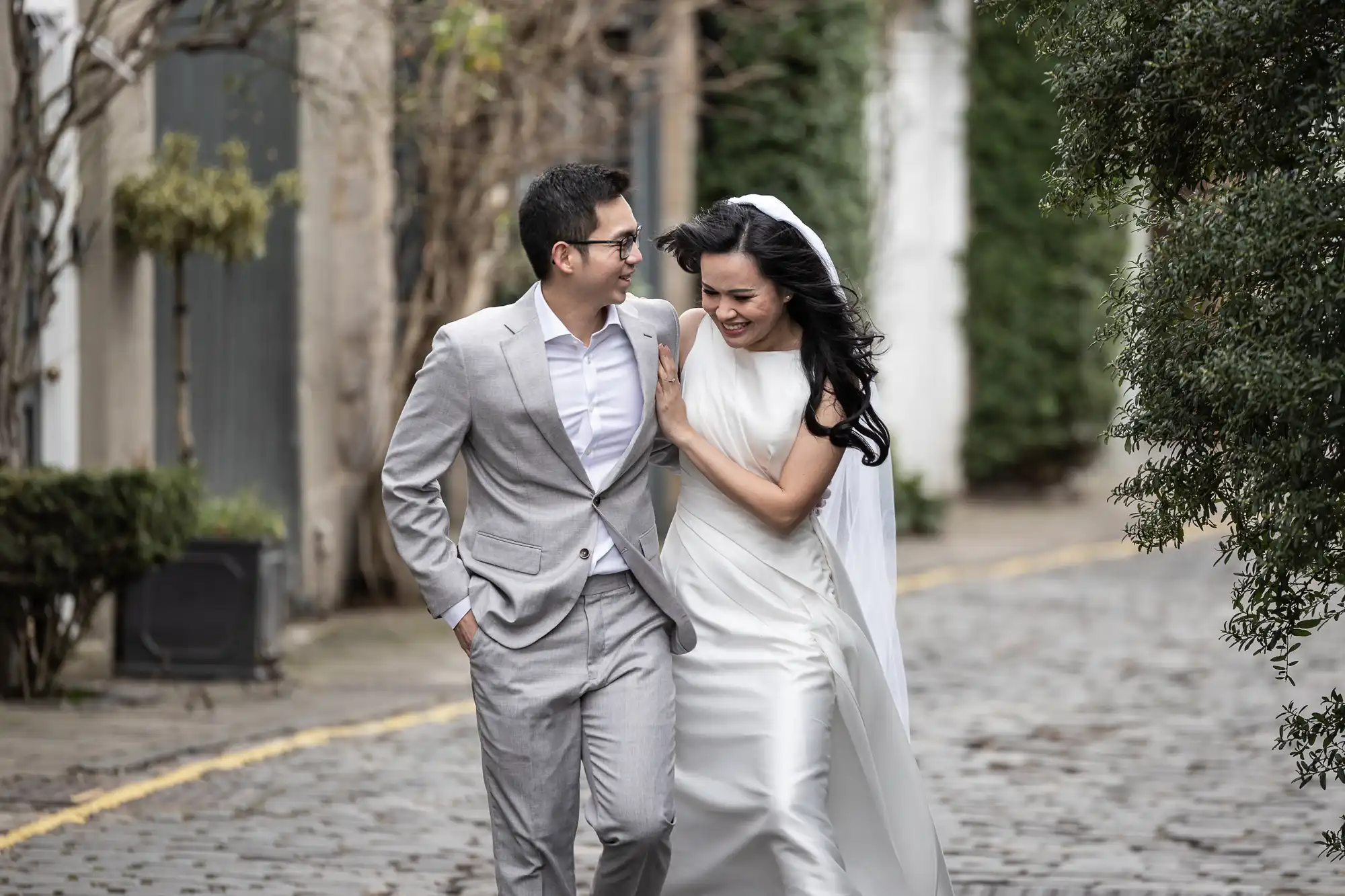 A bride and groom walk arm in arm along a cobblestone path, the groom in a light gray suit and the bride in a white dress with a veil.