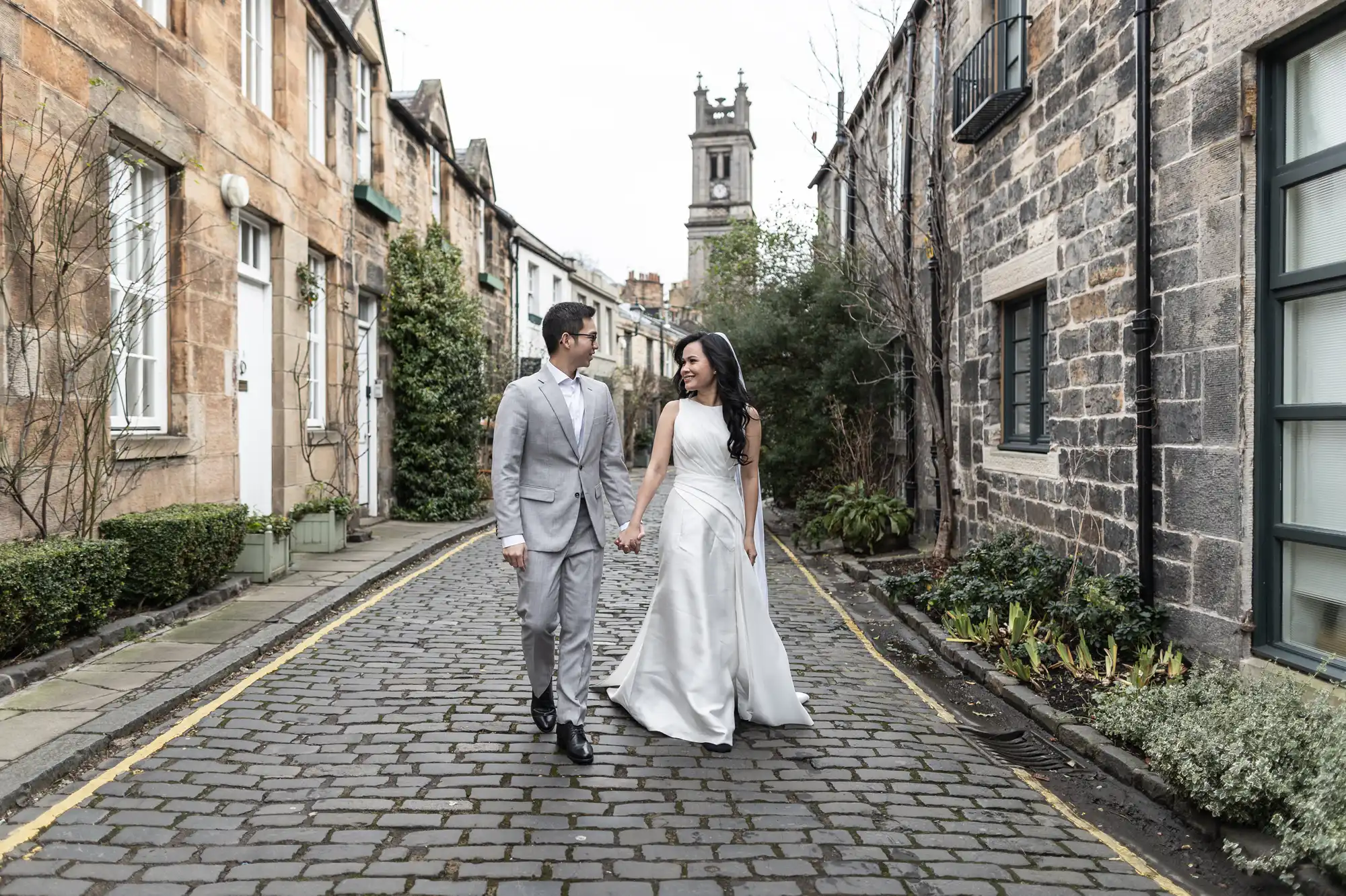 A couple in wedding attire walks hand in hand down a cobblestone street lined with stone buildings and greenery.