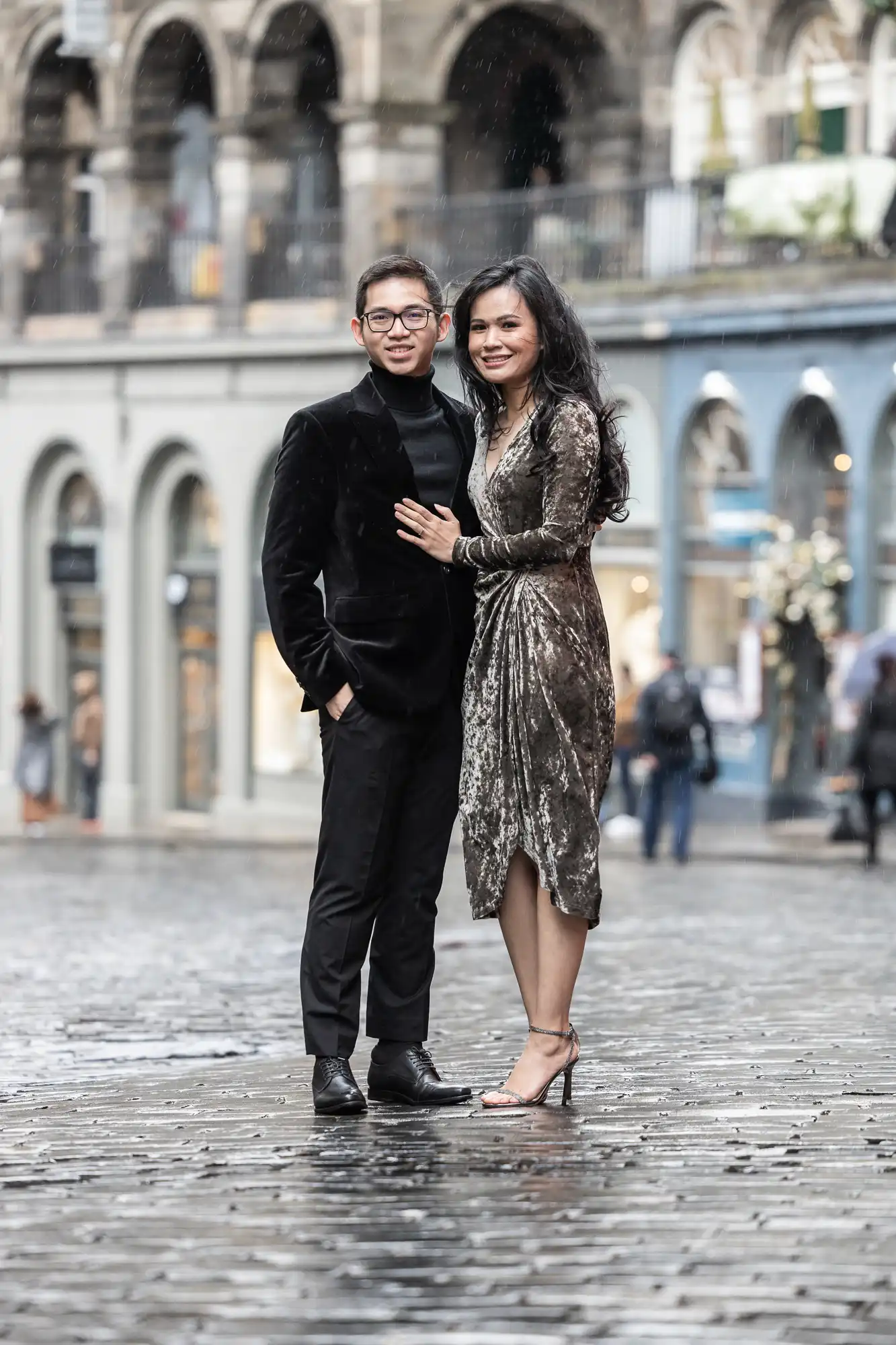 A man and a woman wearing formal attire pose together on a wet cobblestone street in front of arched buildings.