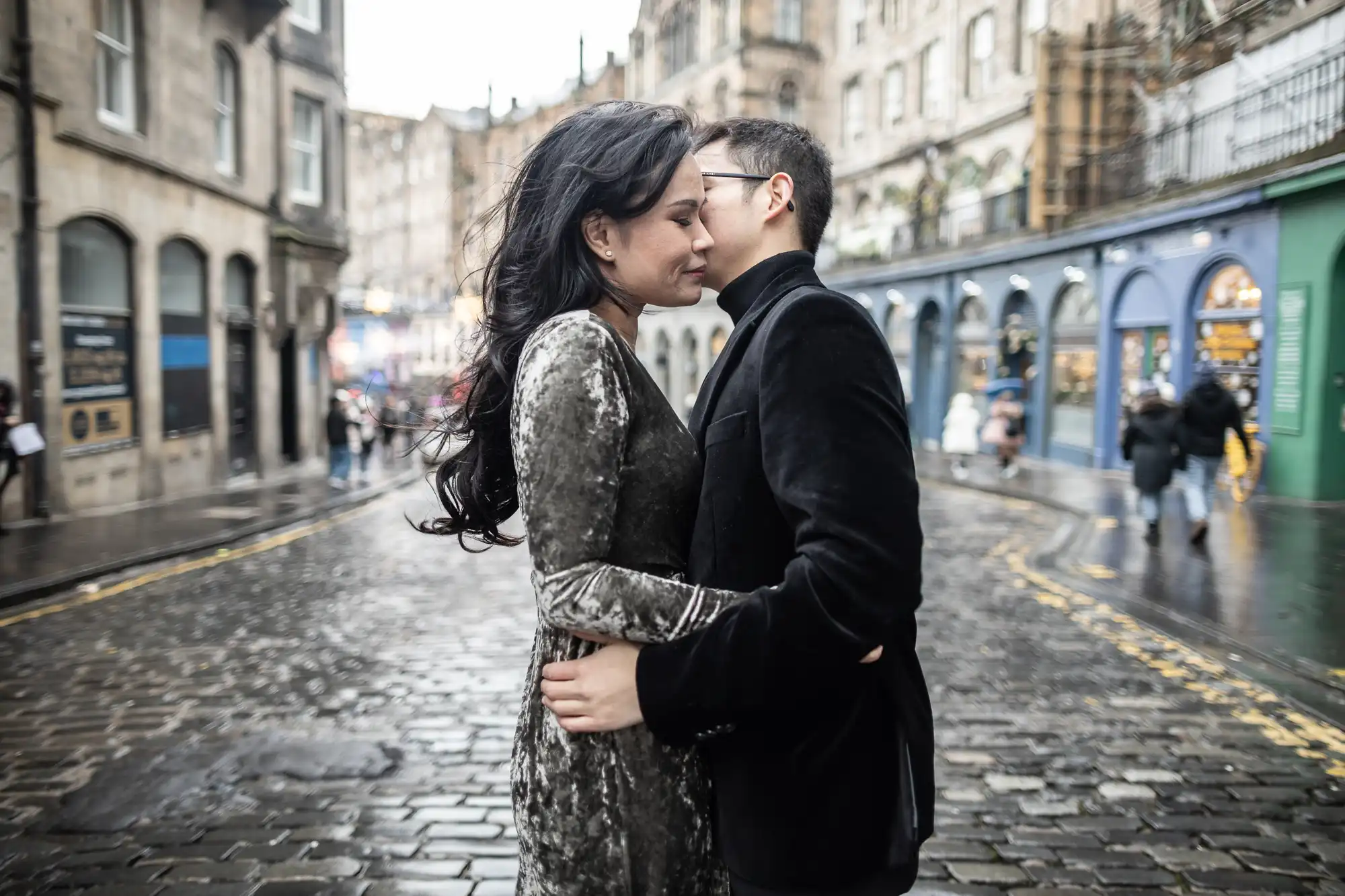 A couple stands close together on a wet cobblestone street in an urban area, surrounded by old buildings with various colorful storefronts. It appears to be an overcast day.