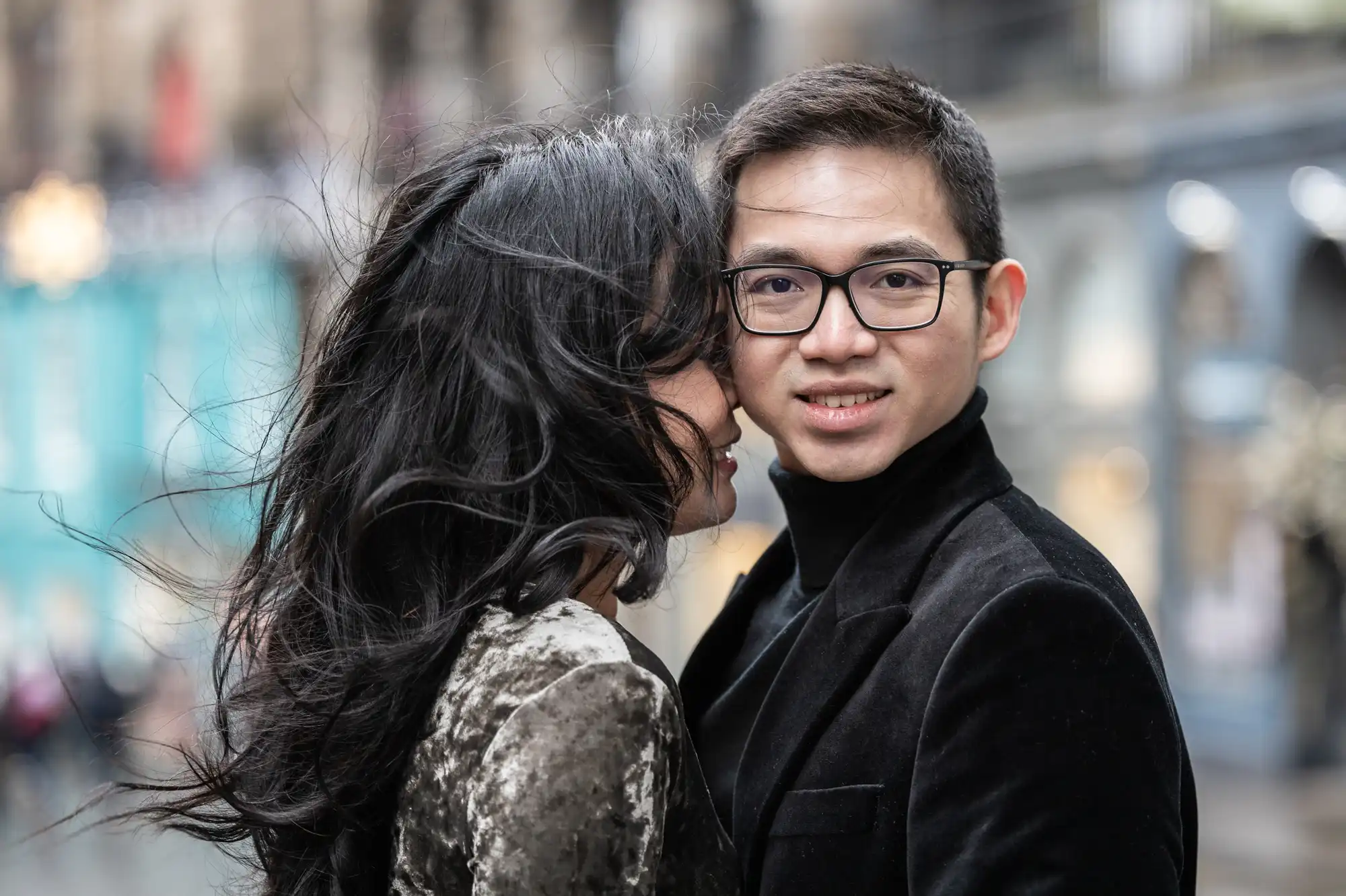 A couple poses outdoors; the woman with long dark hair is leaning towards a man with glasses.