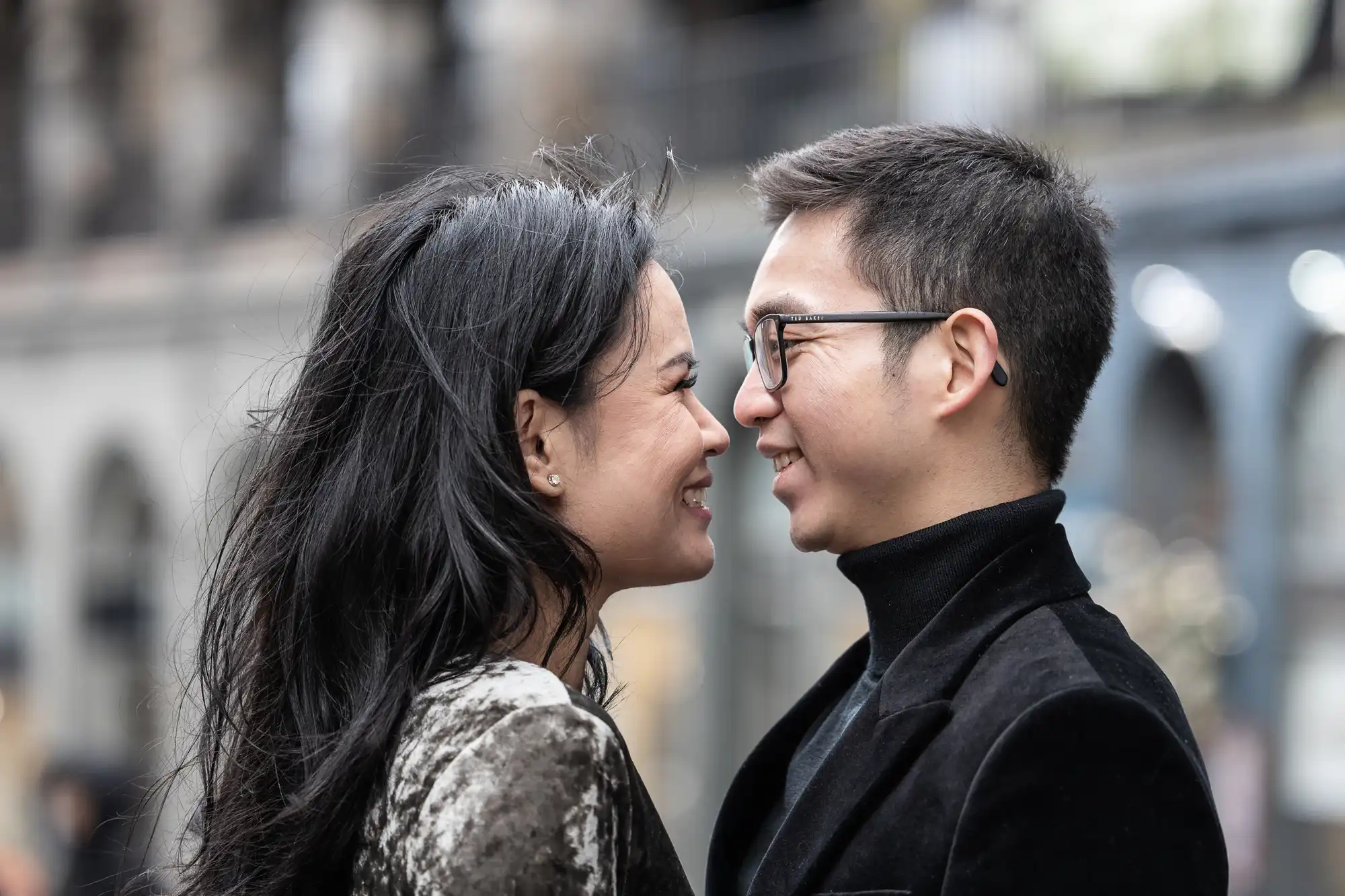 Two people are standing close, facing each other, and smiling. They appear to be outdoors.