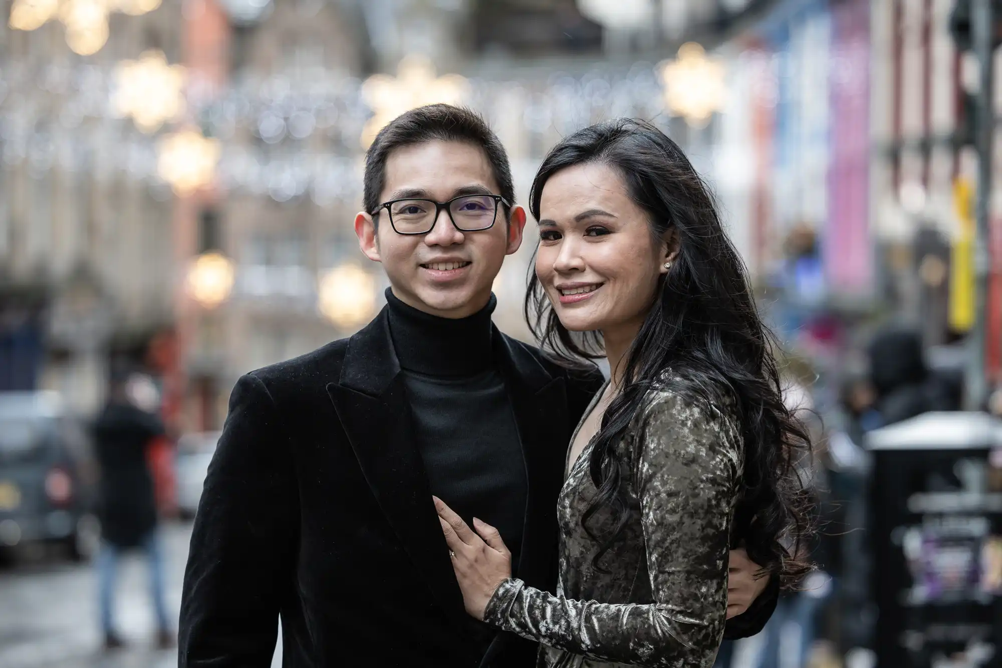 A man and woman stand closely together, smiling at the camera with a street in the background adorned with string lights.