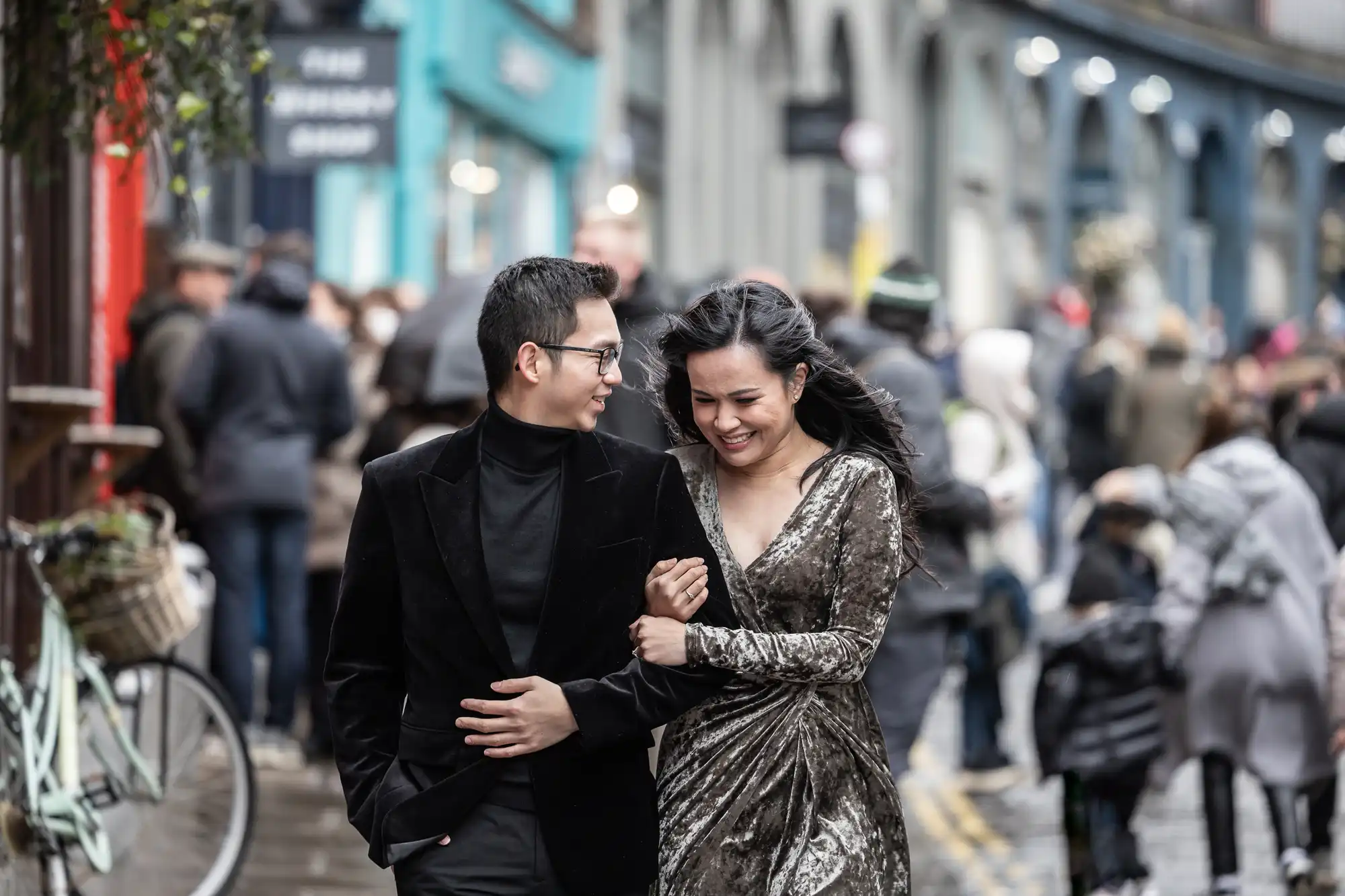 A couple, dressed formally, walks arm-in-arm down a crowded street with shops and people in the background.