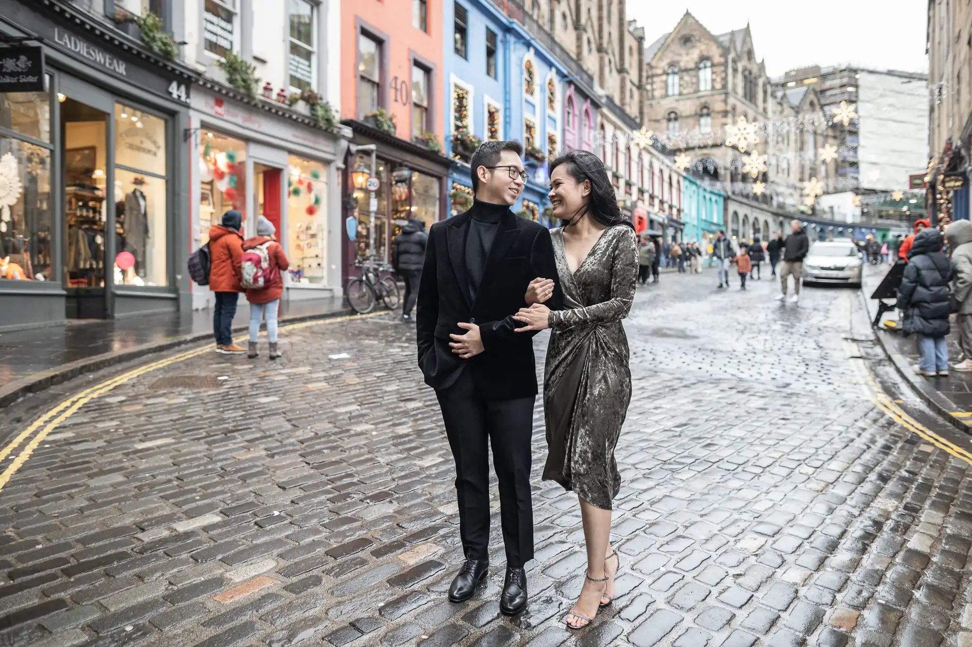 A well-dressed couple walks arm-in-arm along a cobblestone street lined with colorful buildings and holiday decorations.