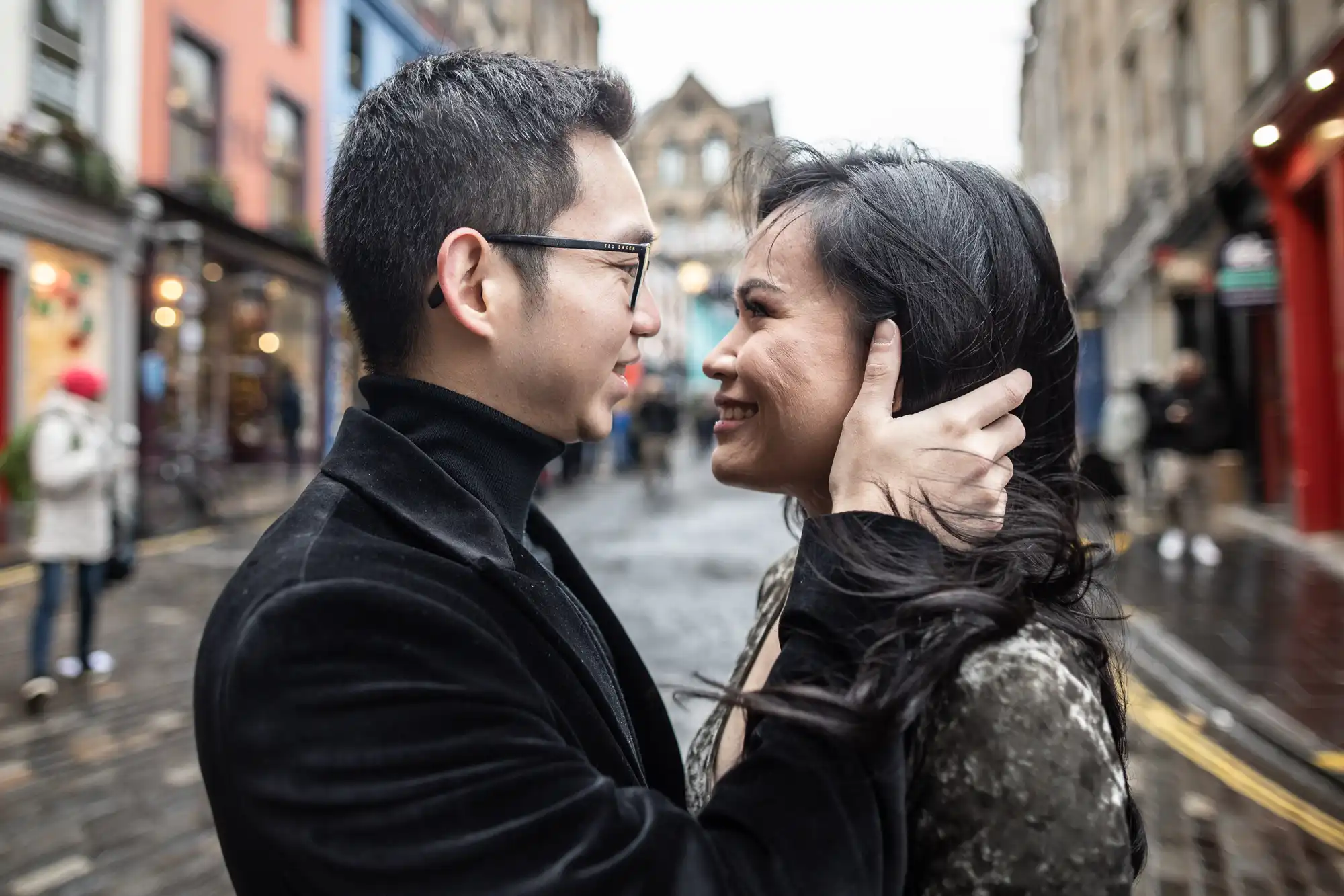 A man and woman stand closely on a narrow street, looking at each other warmly as the man gently holds the woman's head. Urban buildings and pedestrians are visible in the background.