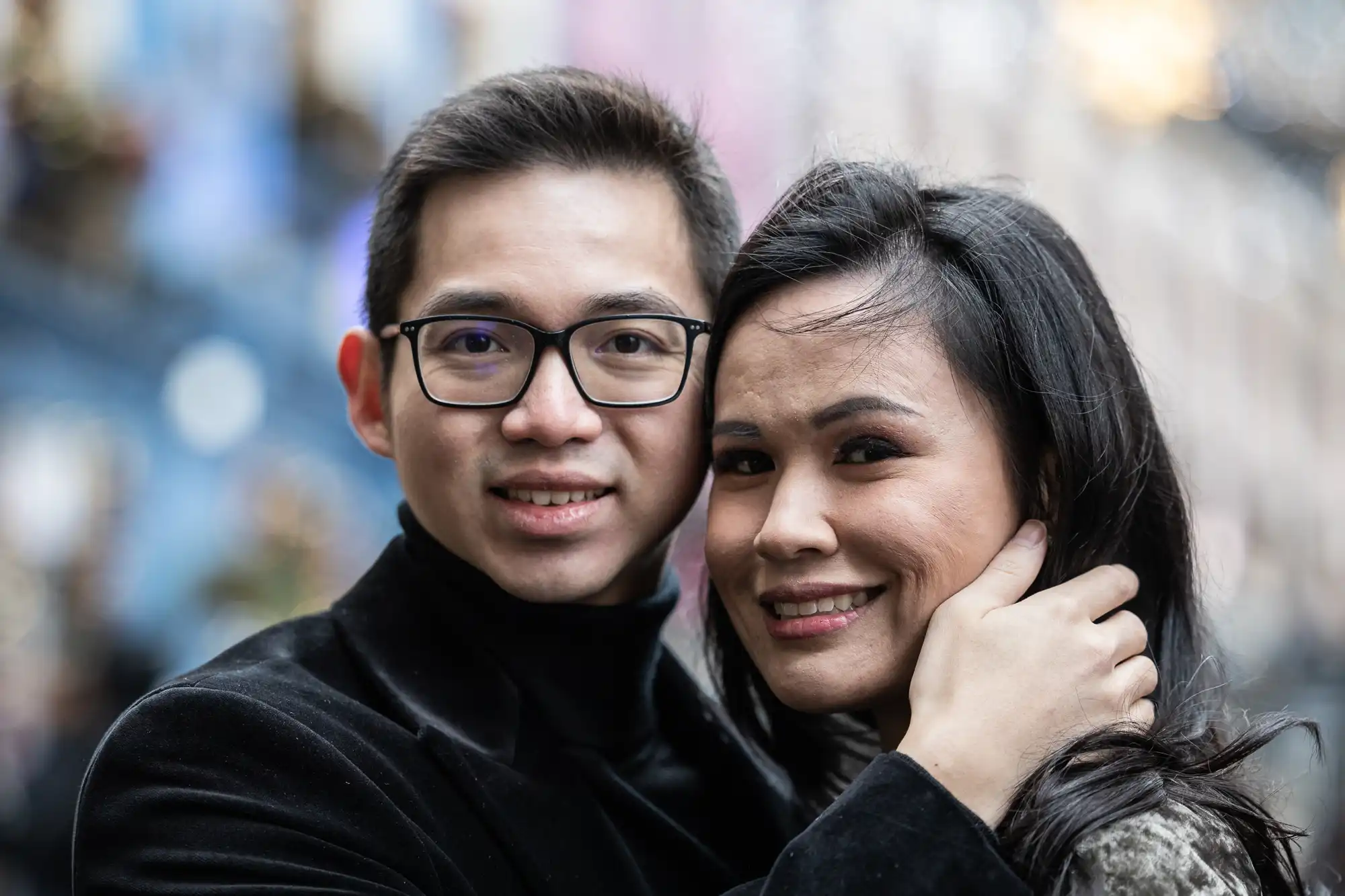 A man and woman smile at the camera; the man is wearing glasses and a dark jacket, and the woman is being held gently by him with a blurred background.