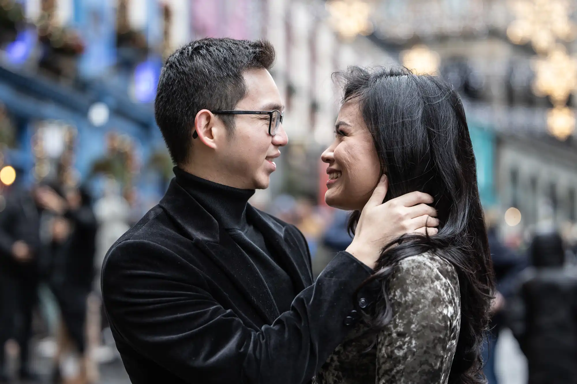 A man and woman smiling at each other while the man gently holds the woman's neck. They are outdoors with blurred buildings and lights in the background.