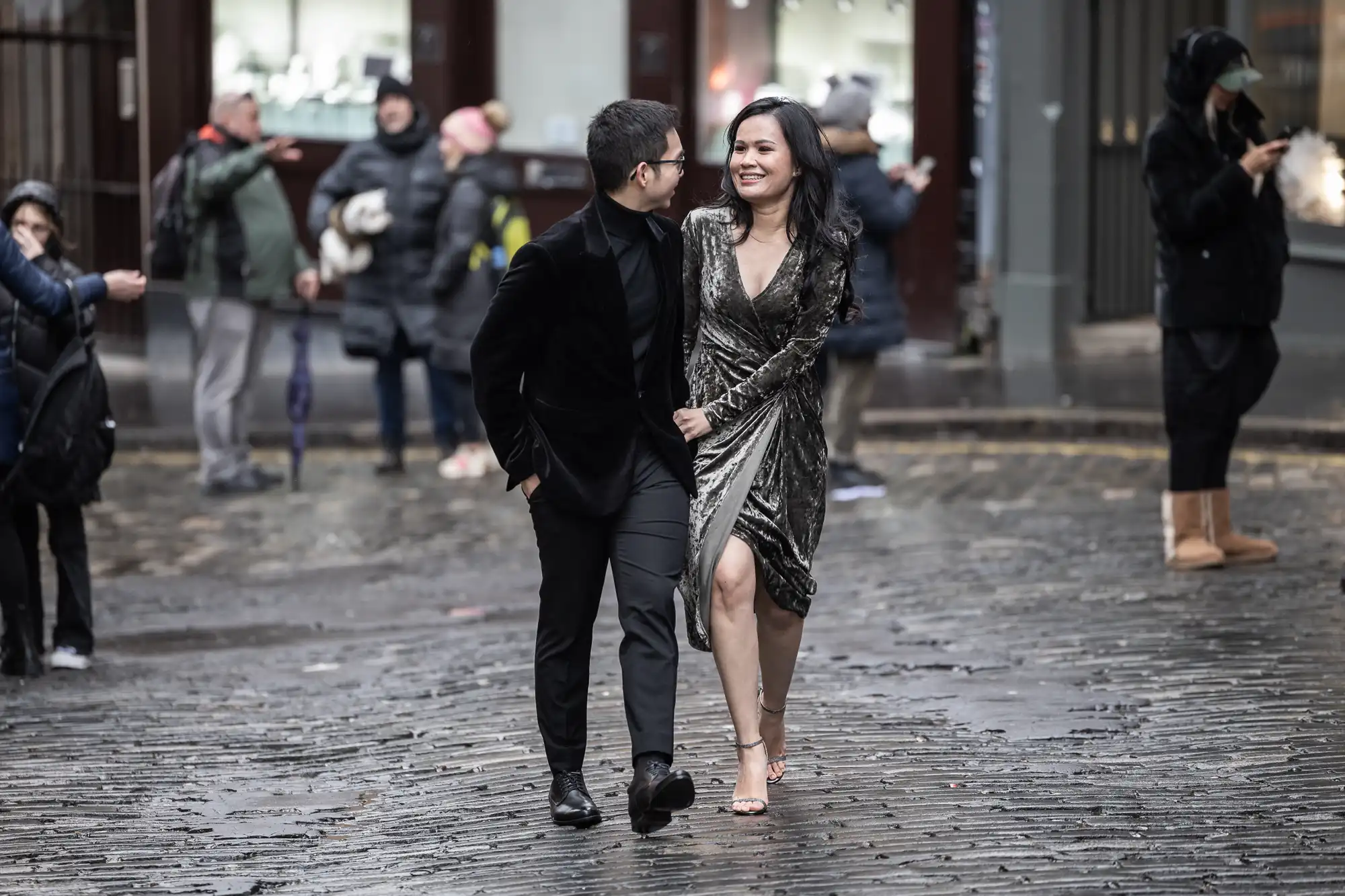 A couple dressed in formal attire walks arm-in-arm on a wet cobblestone street, smiling and looking at each other. People bundled in winter clothing are seen in the background.