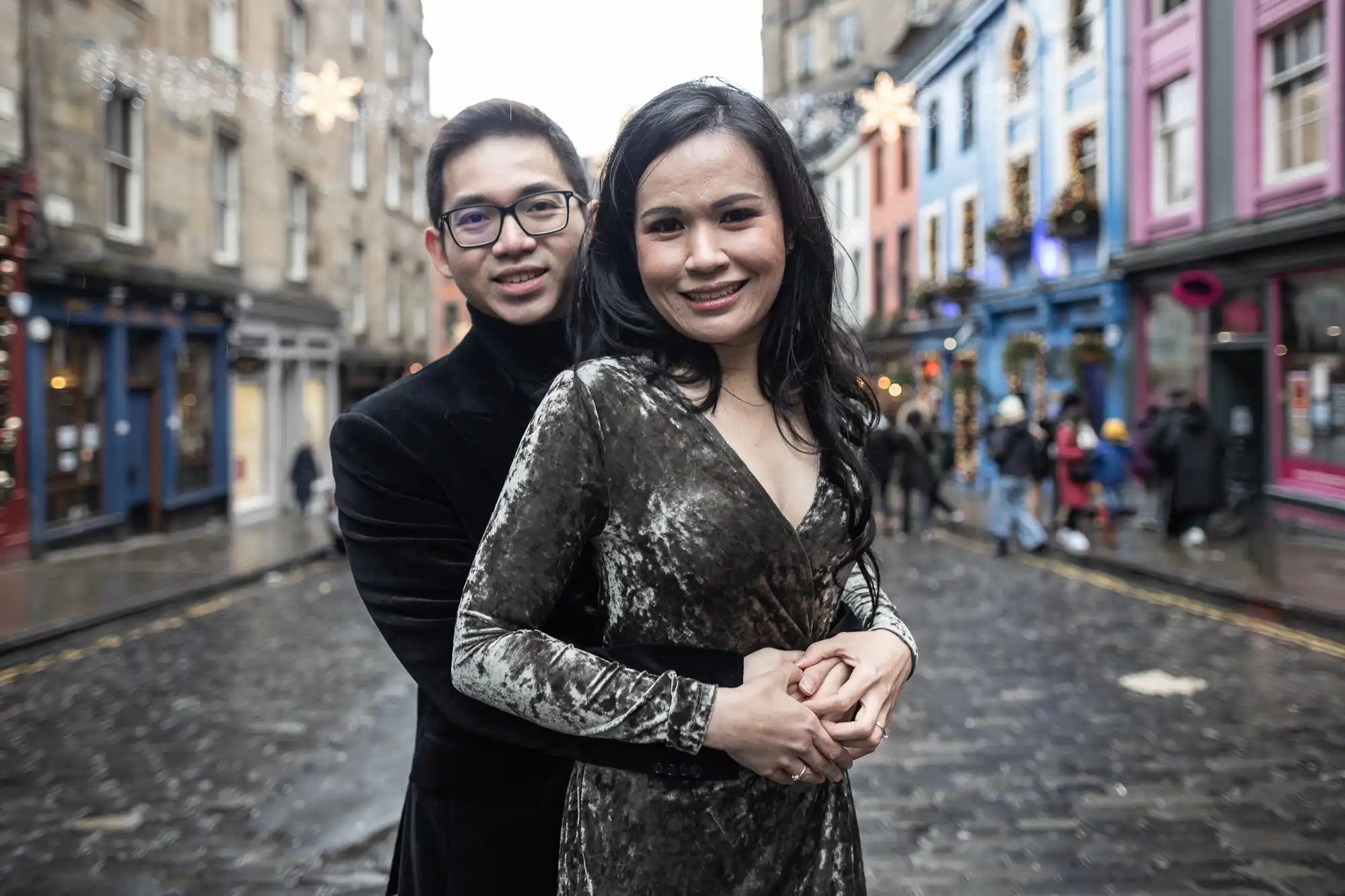 A couple stands close together embracing on a cobblestone street lined with colorful buildings.