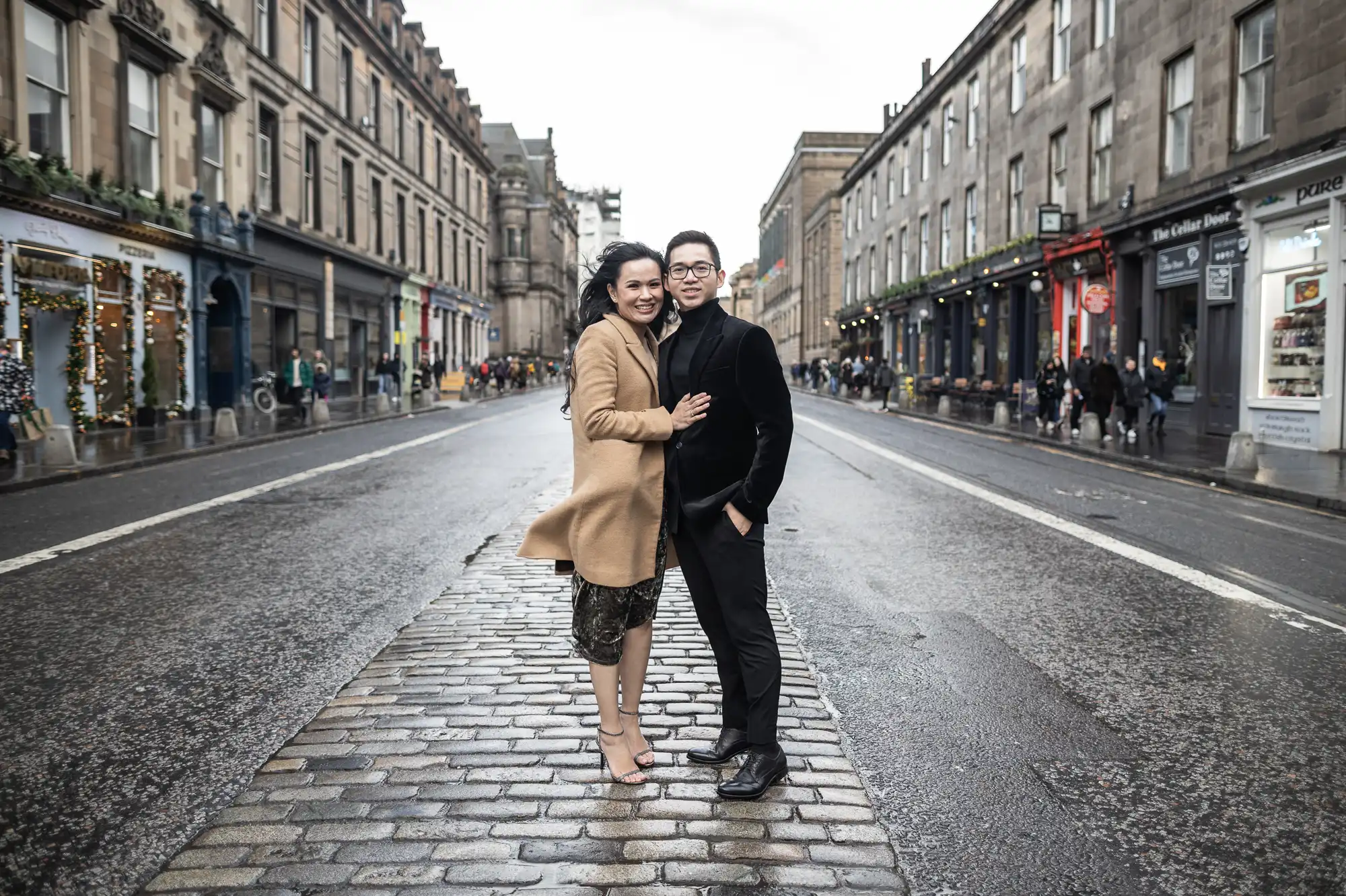 A couple stands together on a wet cobblestone street flanked by buildings and shops, with a crowd and festive decorations in the background.