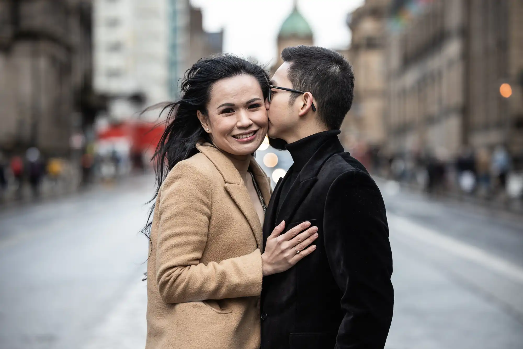 A woman in a tan coat smiles at the camera while a man in a black coat kisses her on the temple. They stand in the middle of a city street.