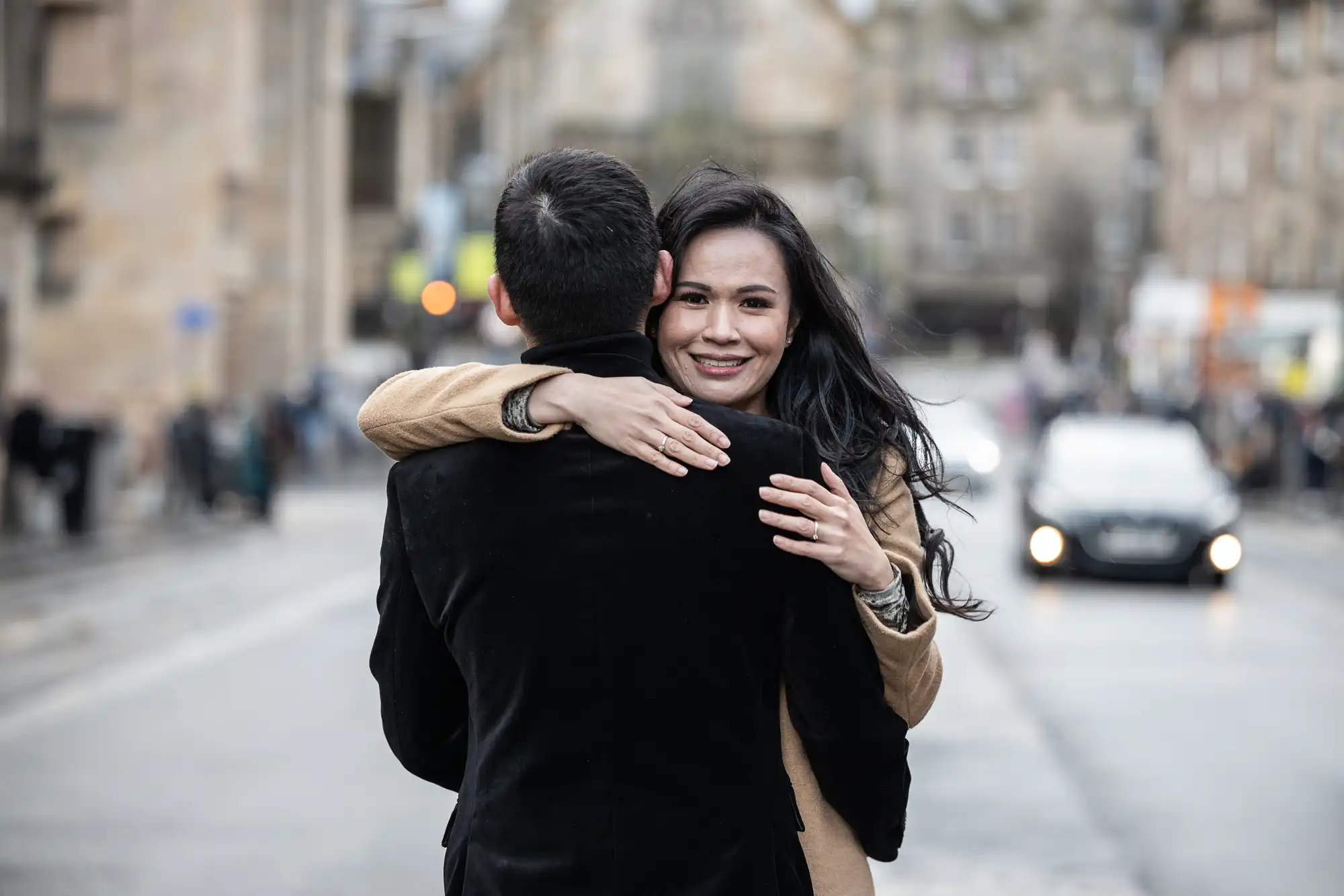 A woman with long dark hair embraces a man on a city street, smiling at the camera. Cars and buildings are visible in the blurred background.