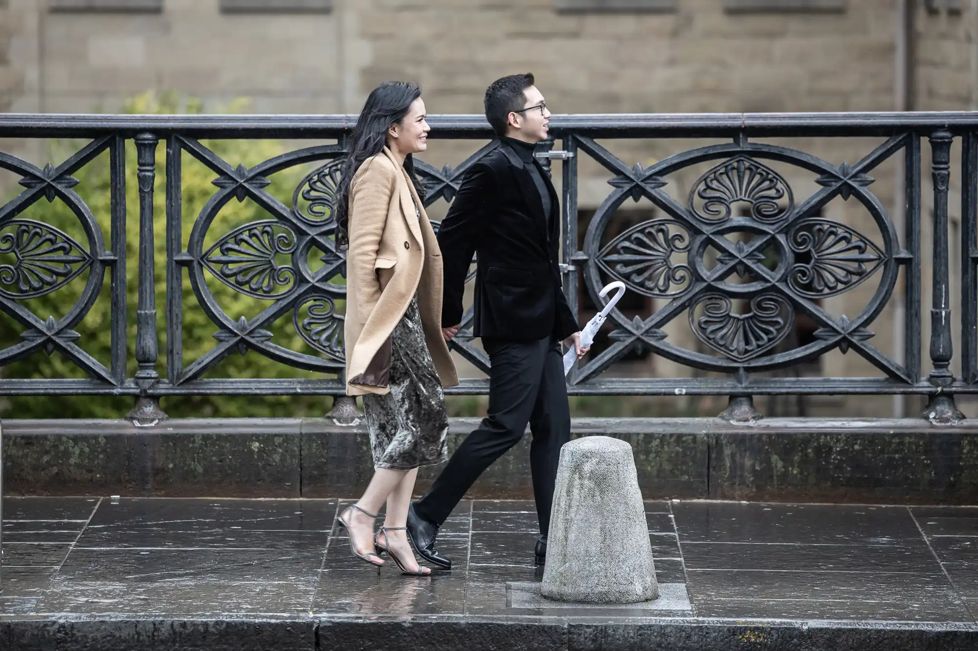 A man and woman walk side by side on a rainy day, dressed in formal attire. The man carries a rolled-up newspaper. They are on a sidewalk with ornate railing behind them.