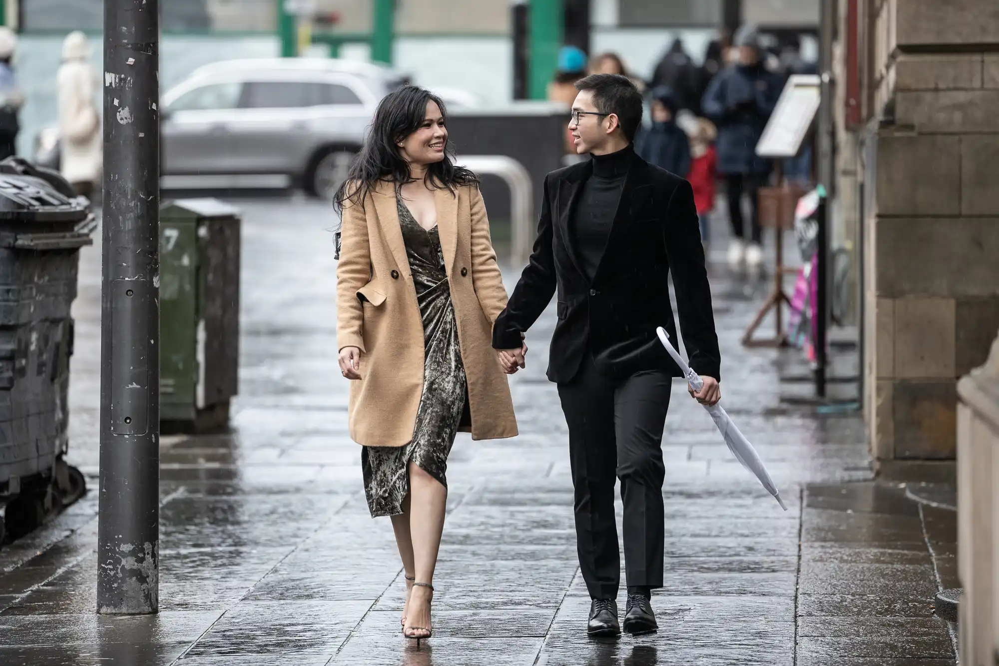 A couple walks hand in hand on a wet sidewalk. She wears a dress and beige coat, while he wears a black suit and holds a closed umbrella.