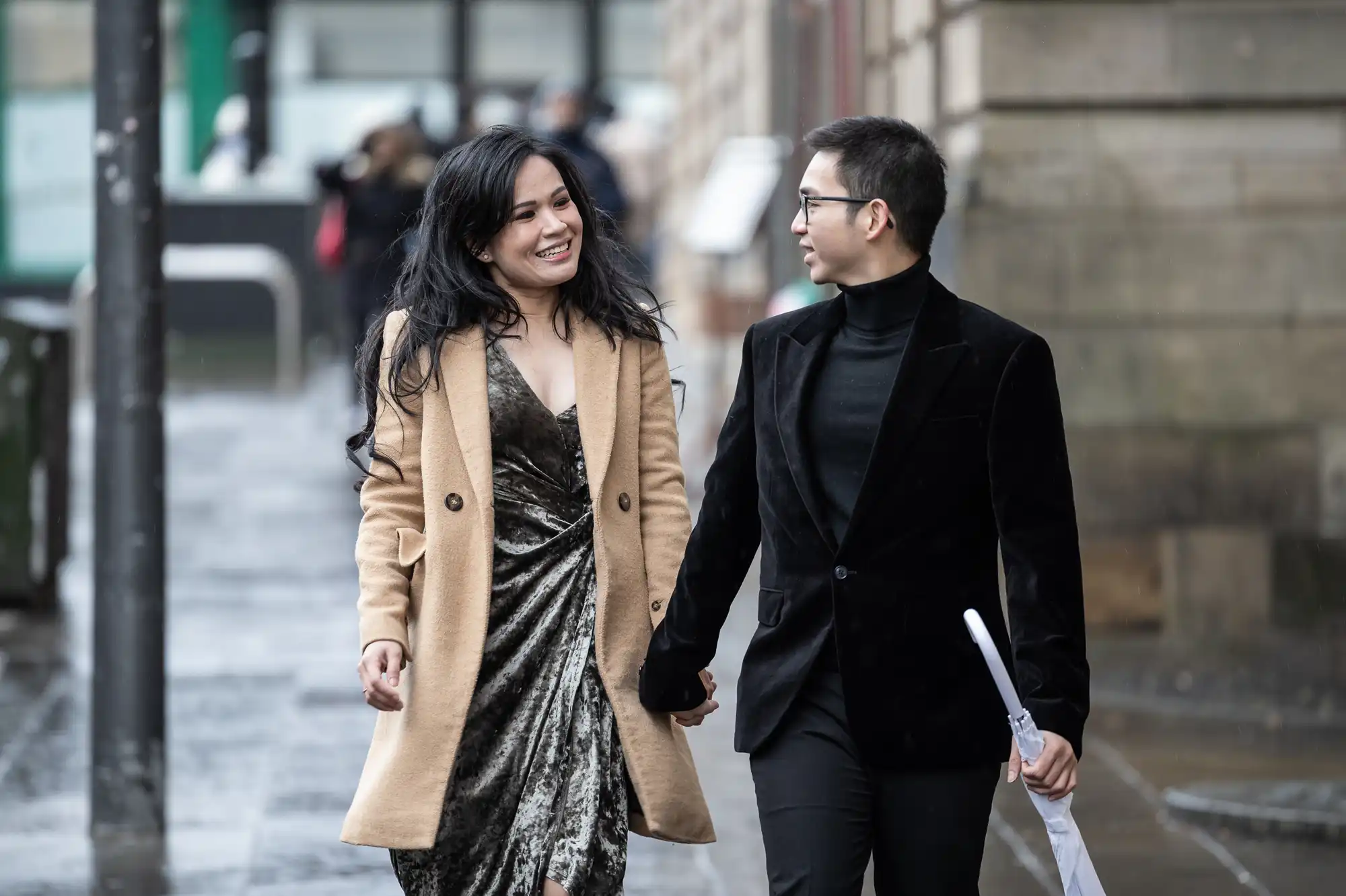 A woman in a brown coat and a man in a black coat walk hand in hand on a rainy street, both smiling in conversation. The man holds an umbrella.