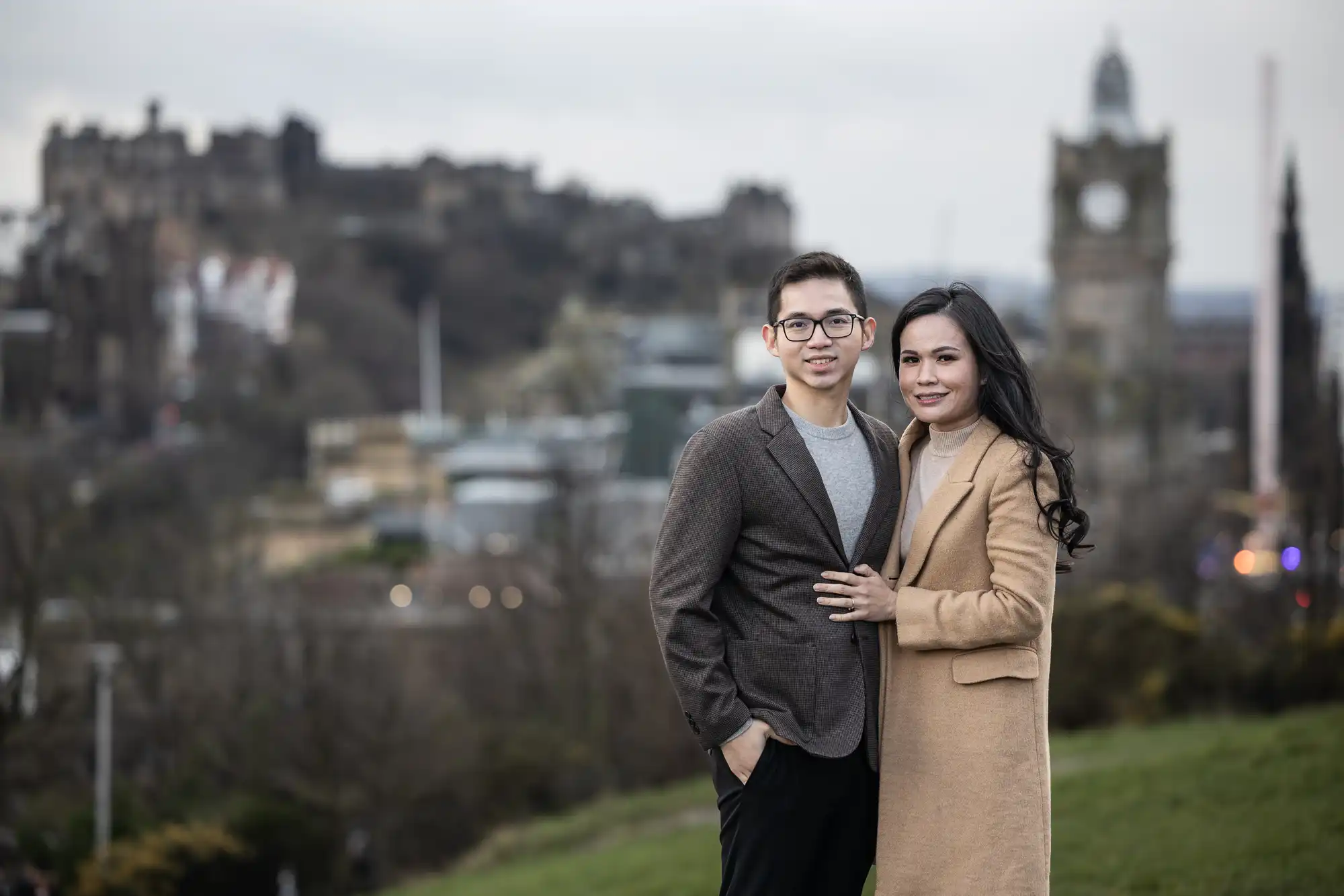 A man and woman stand outdoors on a hillside with a blurred cityscape and historic buildings in the background. Both are wearing coats, with the woman having her hand on the man's chest.