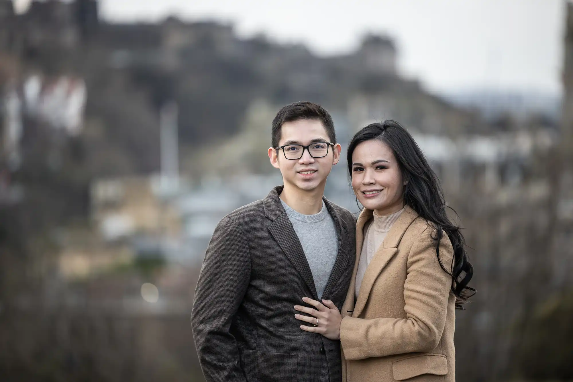 A man and a woman stand outdoors against a blurred natural background. They are both dressed in formal winter clothing and smiling at the camera.