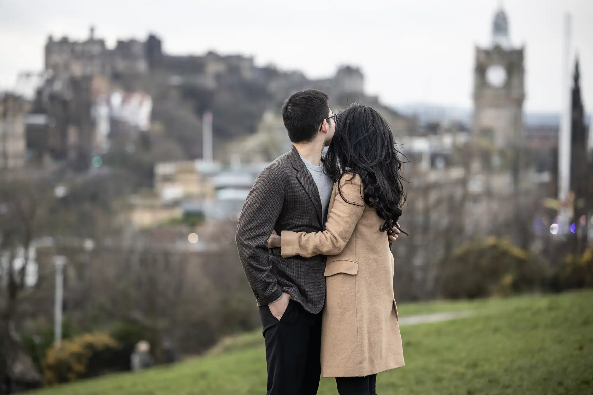 A couple stands on a grassy hill, embracing and looking toward a distant cityscape with historic buildings and a clock tower.