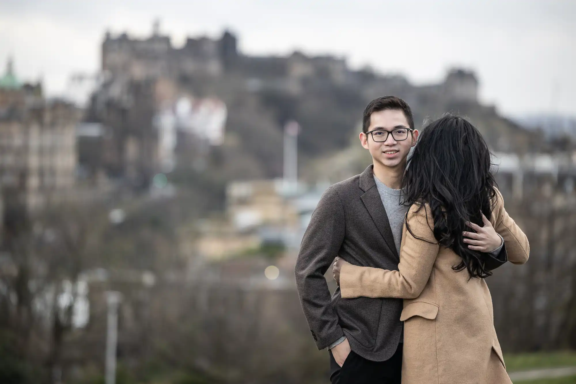 A person wearing glasses and a brown blazer stands while another person with long dark hair and a tan coat hugs them. An out-of-focus cityscape with historic buildings is in the background.