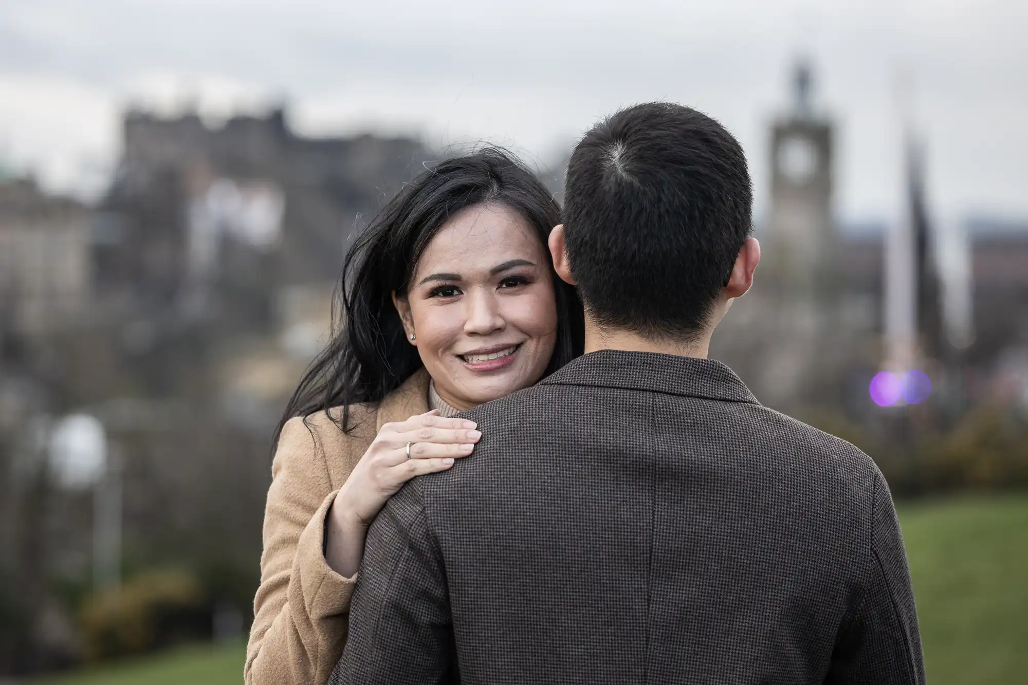 A woman smiling, embracing a man facing away, with a blurred outdoor cityscape in the background.