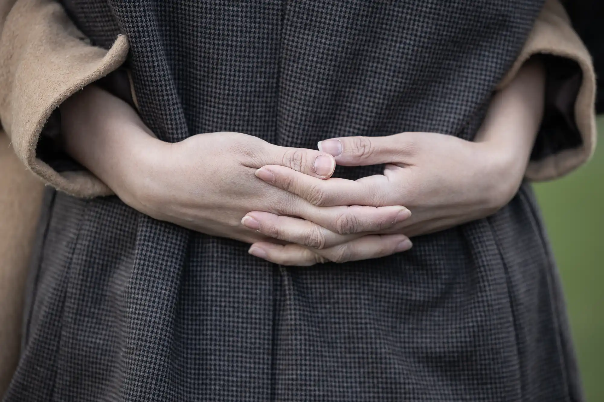 Person wearing a dark checkered coat, with hands clasped together in front.