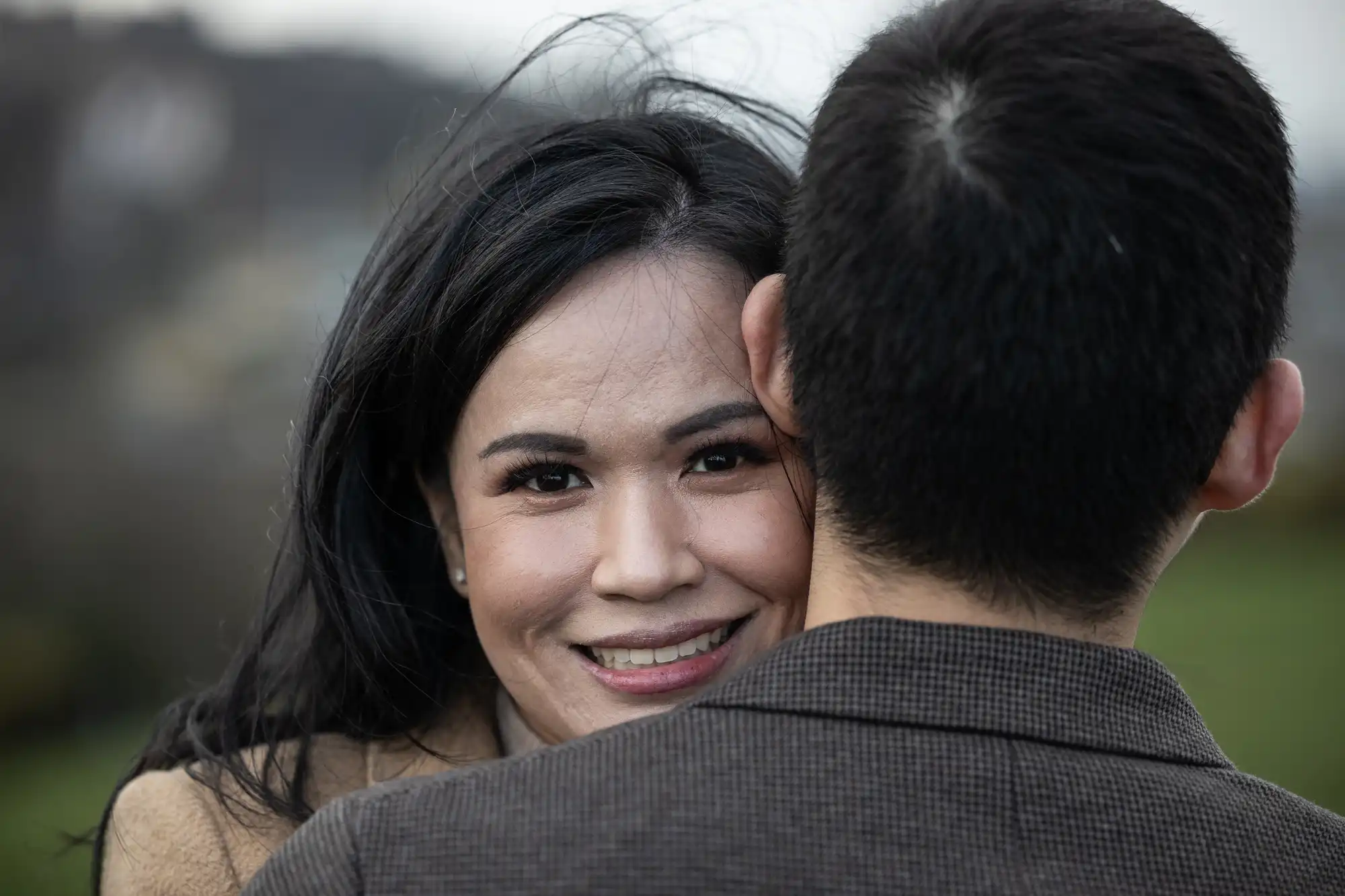 A woman smiles at the camera while embracing a man with her arms around him. The man's back is turned towards the camera. Both are outside.