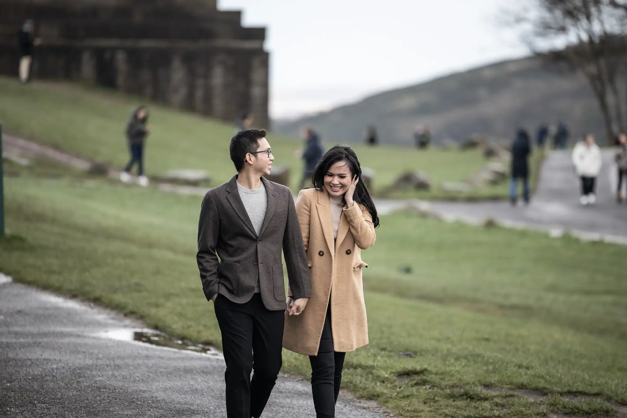 A couple walks hand in hand on a paved path in a park, with people in the background and a hill visible in the distance. The man is wearing a brown jacket, and the woman is wearing a beige coat.