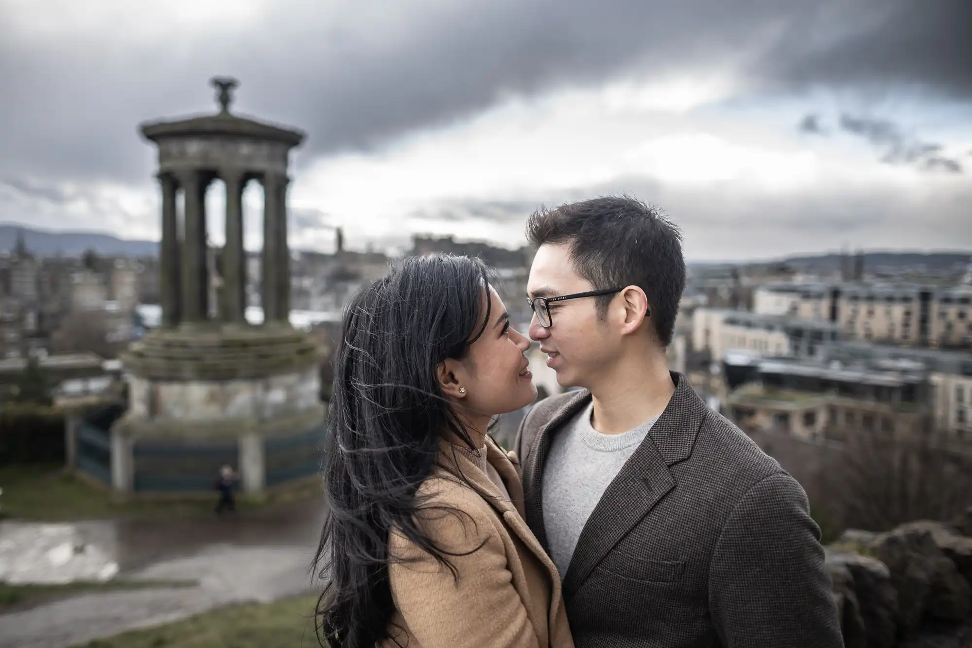 A couple stands close together, gazing at each other, with a historical monument and cityscape in the background under a cloudy sky.