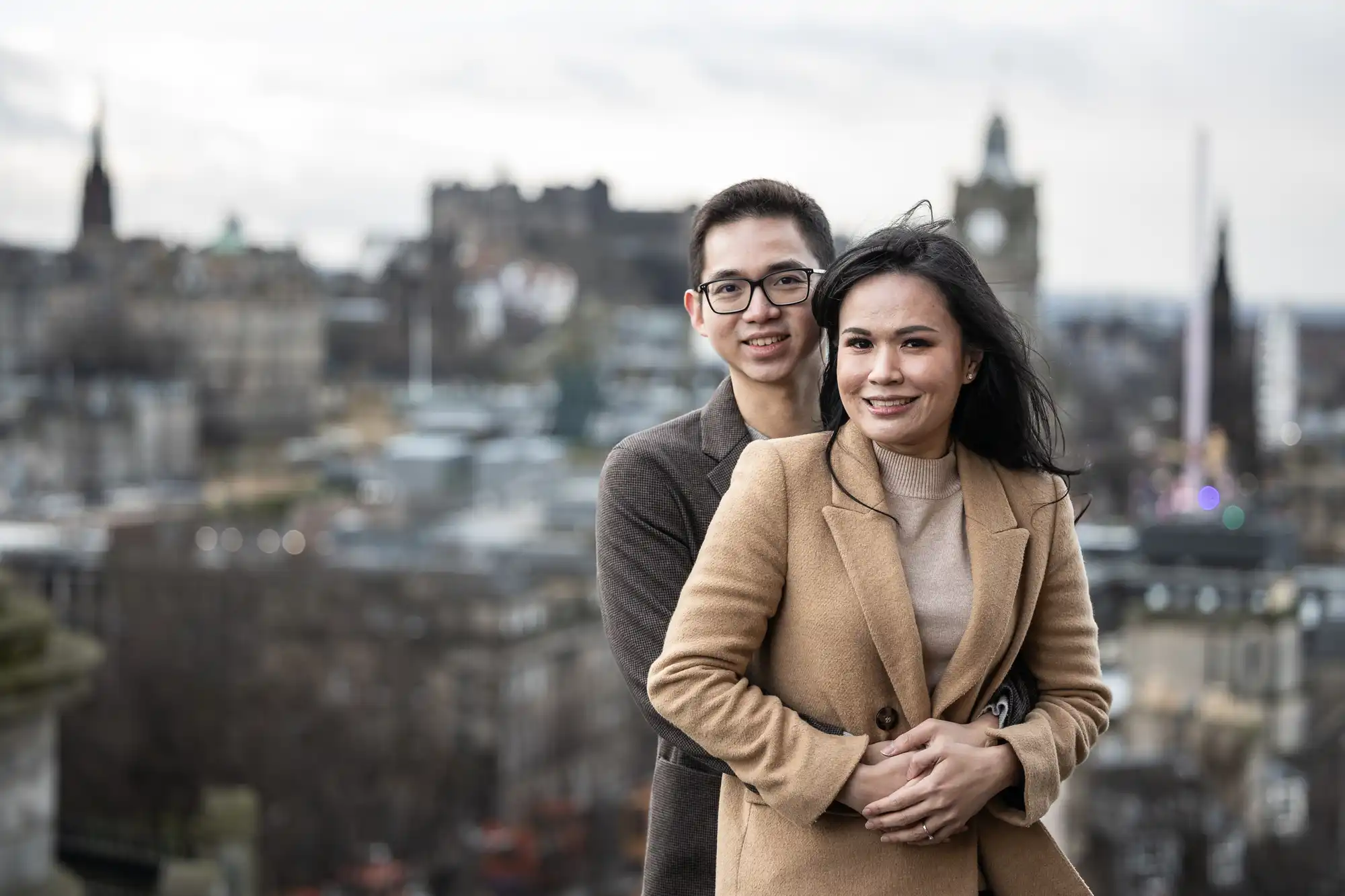 A couple stands together in an outdoor setting with a cityscape in the background. The woman is in front, smiling, while the man stands behind her, holding her around the waist.