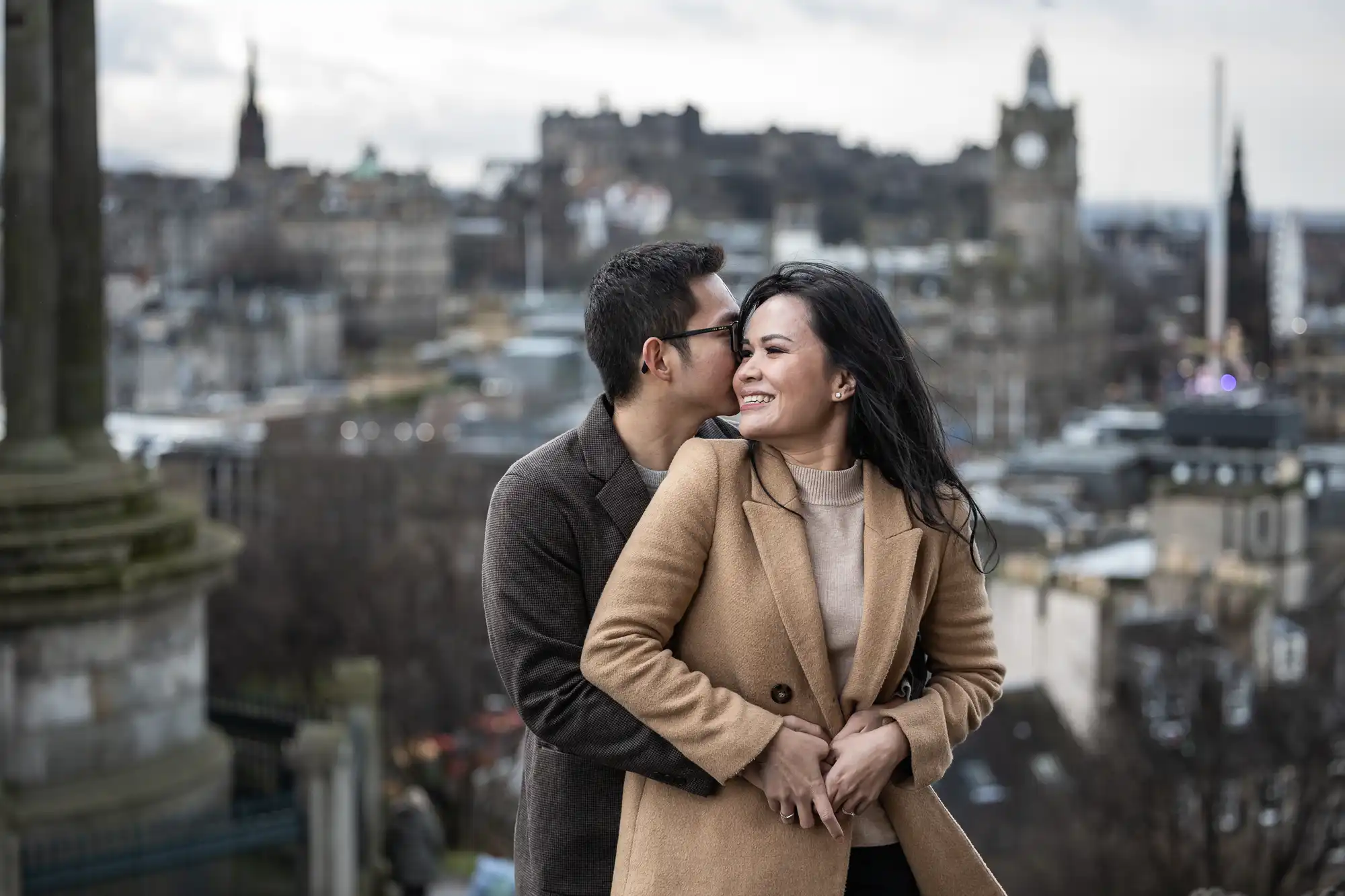 A couple stands close together, the man embracing the woman from behind, with a cityscape in the background.