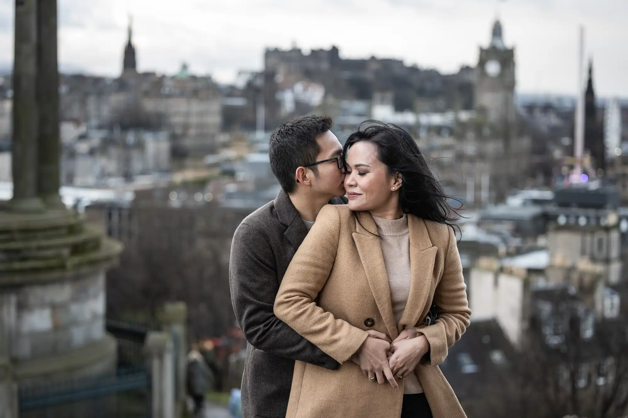 A couple in coats stands on a viewpoint overlooking a cityscape. The man embraces the woman from behind, both smiling with city buildings and a clock tower visible in the background.