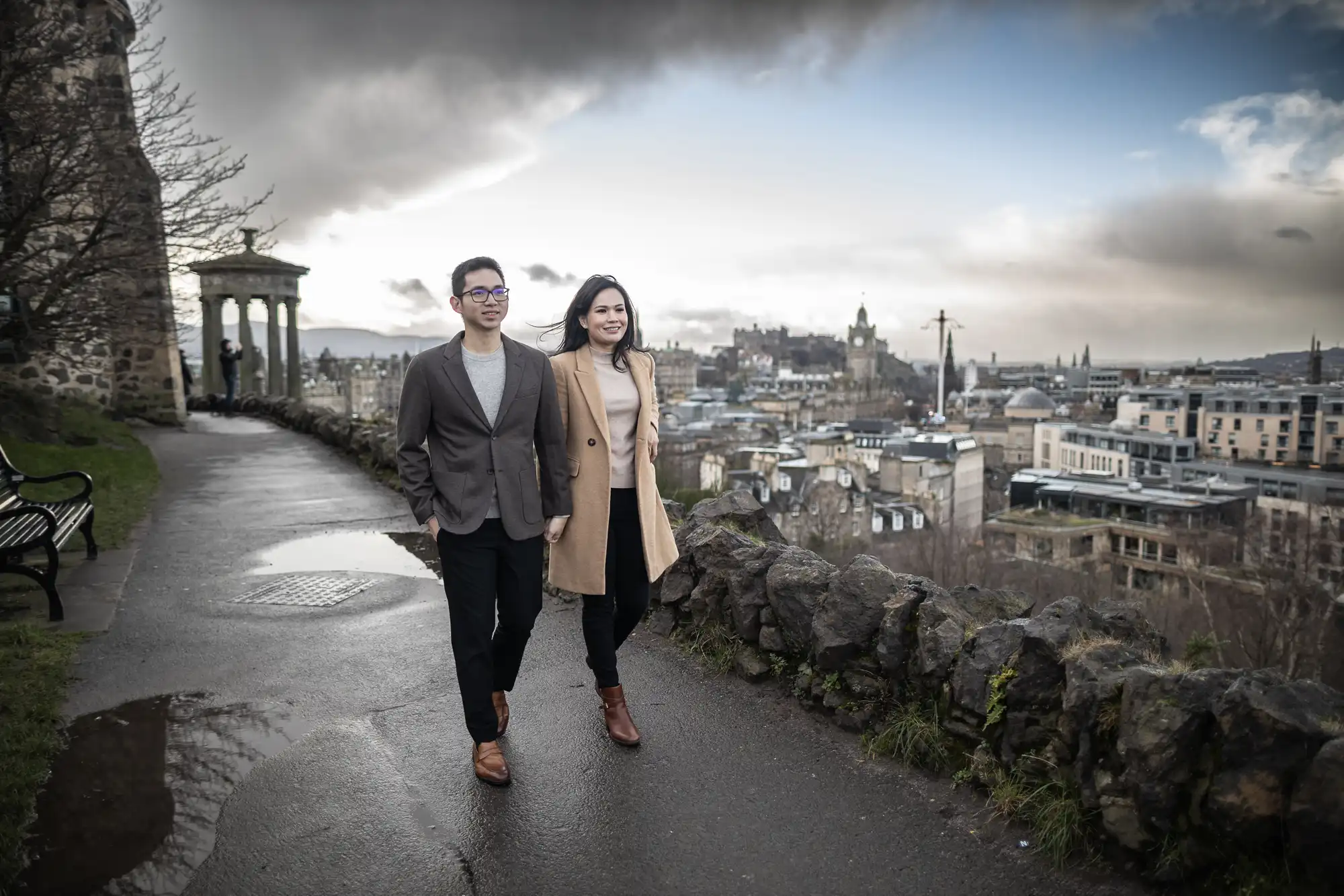 A man and a woman walk along a pathway on a cloudy day, with a cityscape and historical buildings in the background.