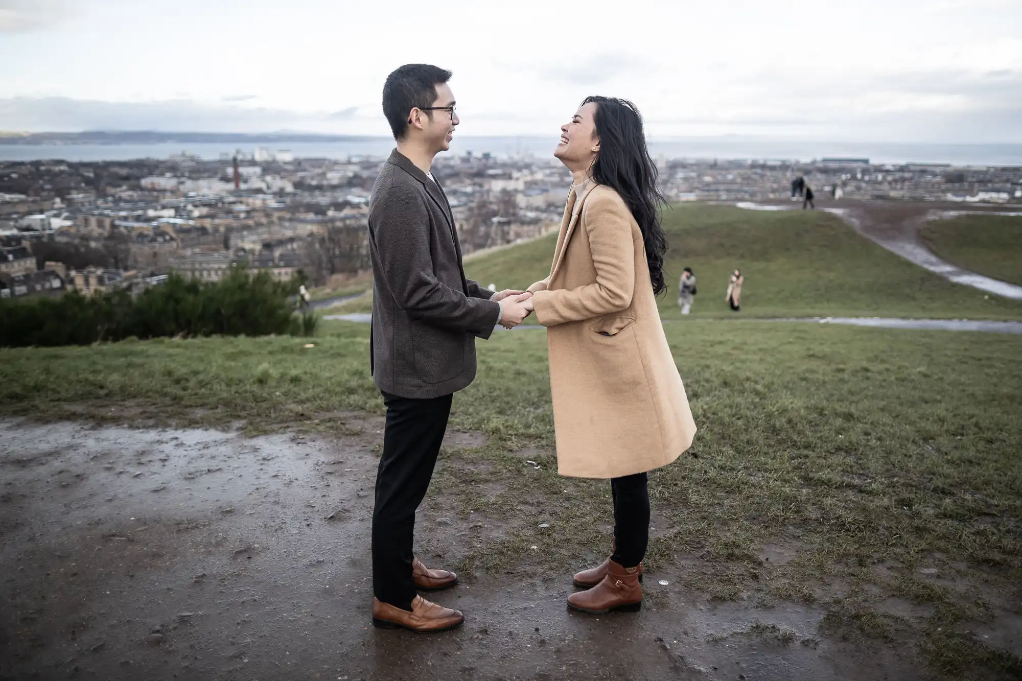 Two people standing on a hilltop holding hands and smiling at each other, with a cityscape in the background. One is wearing a brown coat, and the other is in a dark jacket and black pants.