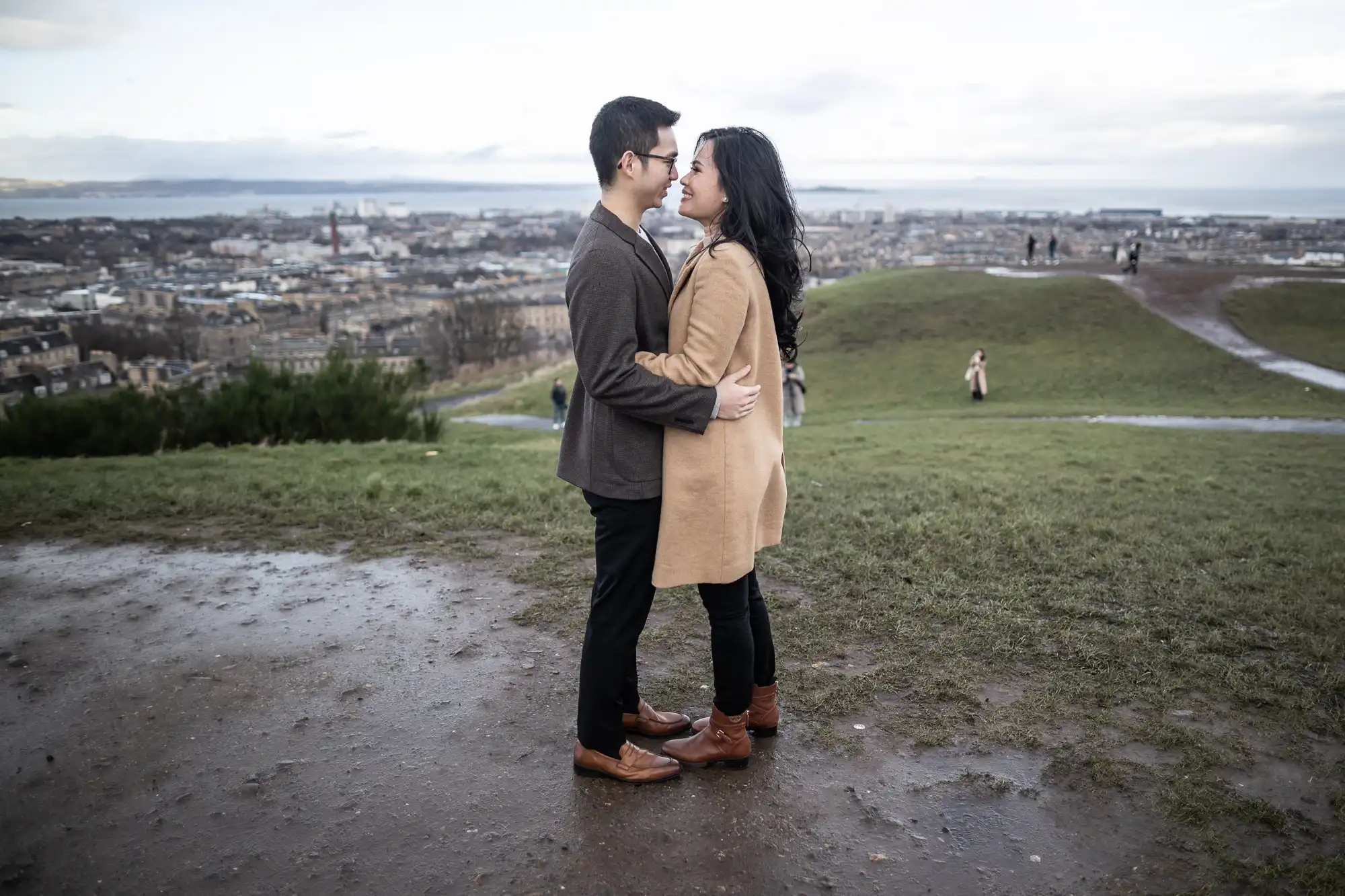 A couple stands facing each other and embracing on a hilltop with a panoramic view of a cityscape in the background. Other people can be seen in the distance on the hill.