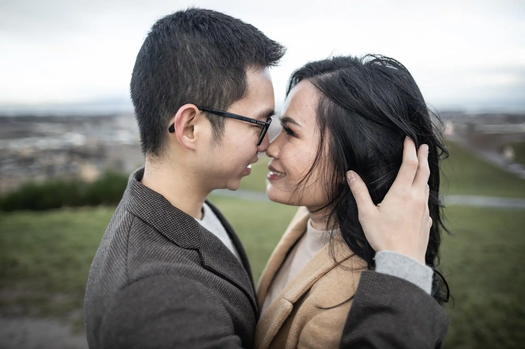 A couple stands close together outdoors, touching foreheads and smiling, with the man holding the woman's head. The background shows a blurred landscape.