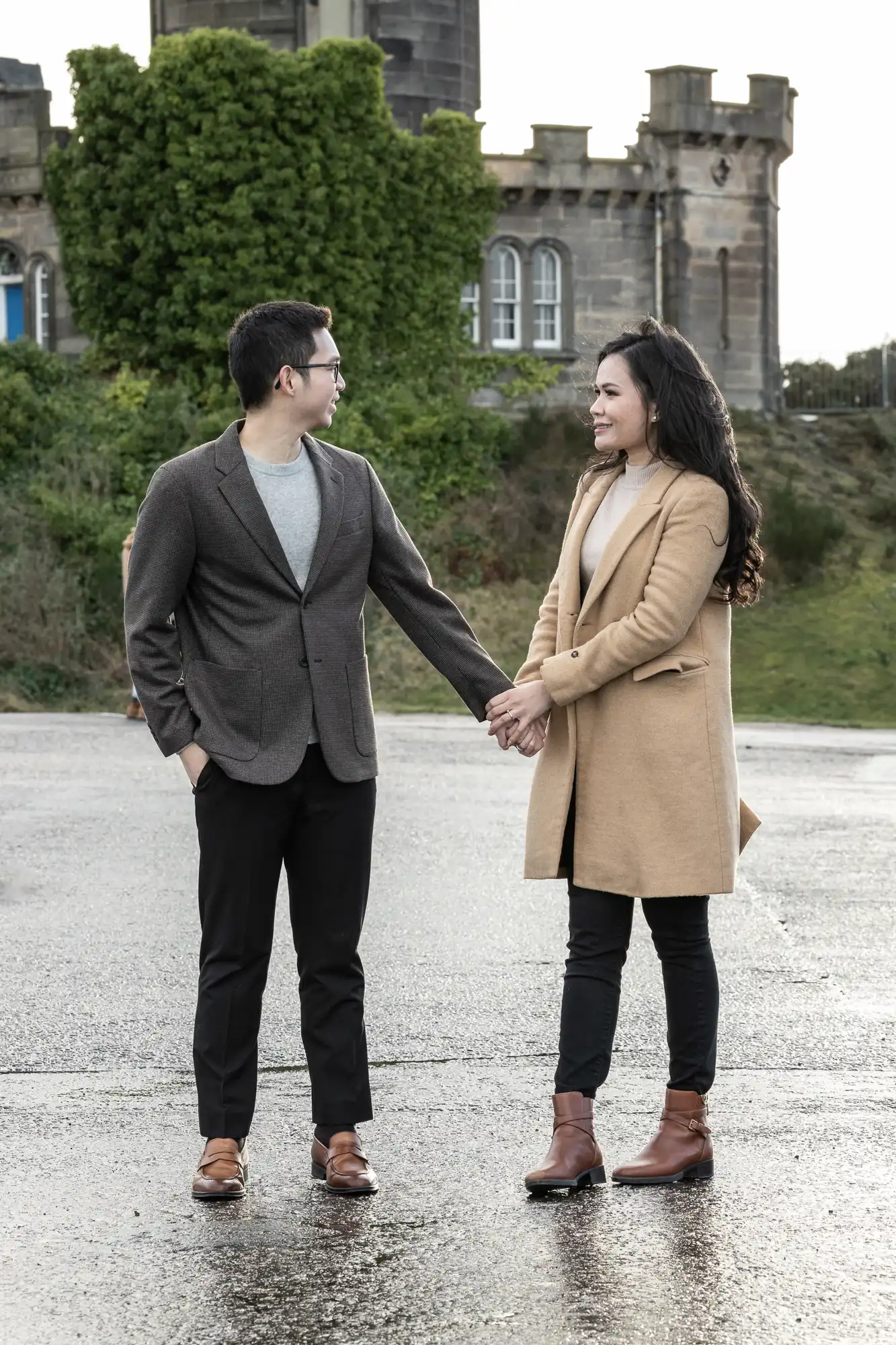 A man and woman stand outside holding hands, both dressed in coats and boots. They face each other, smiling. A large ivy-covered building is in the background.