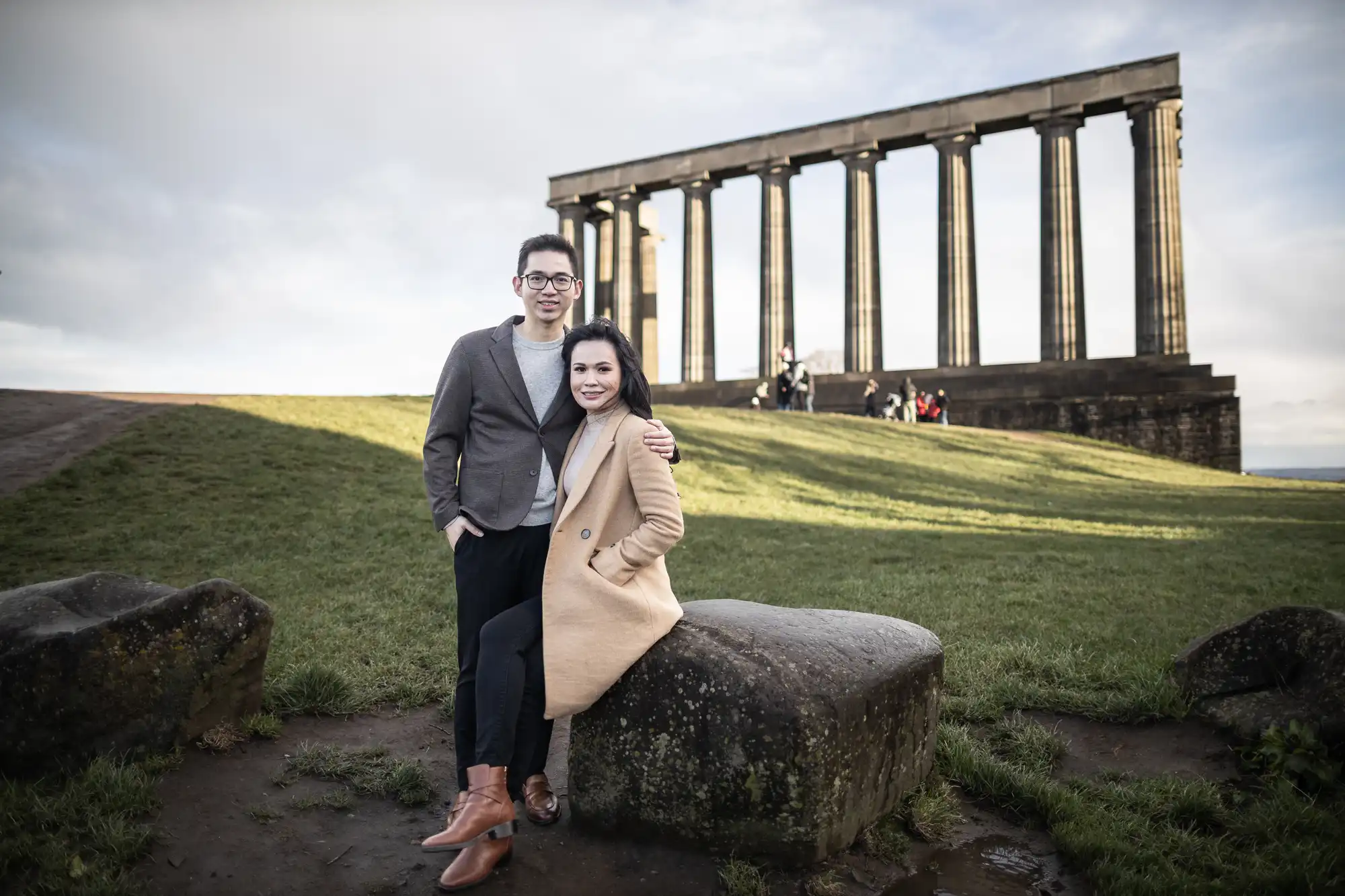 A man and woman pose on a grassy hill in front of a stone structure with pillars. The woman sits on a large rock while the man stands beside her with his arm around her.