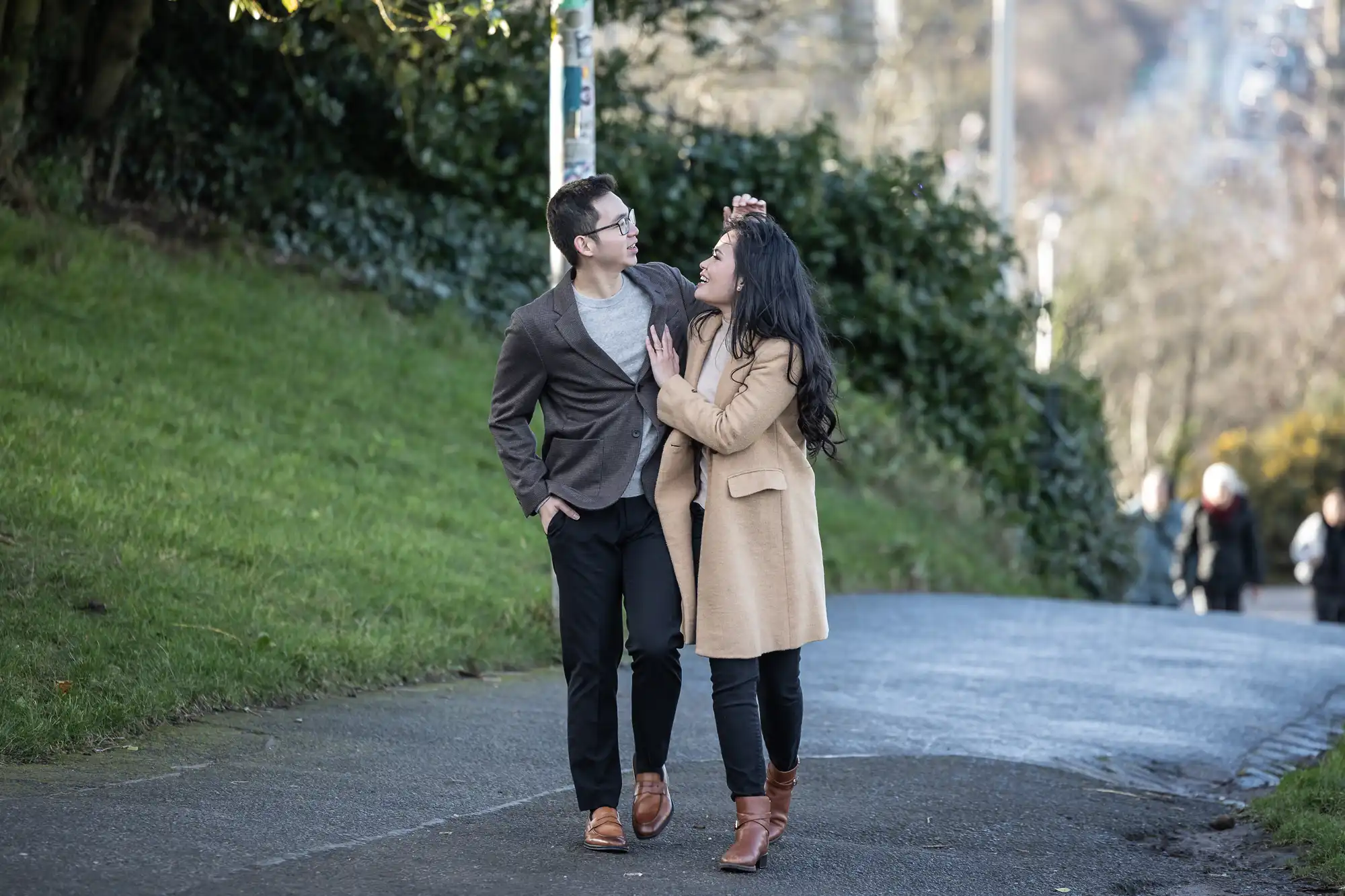 A man and woman walk together on a paved path in a park, engaging in conversation. The man has his arm around the woman's shoulder. Green grass and trees line the path.