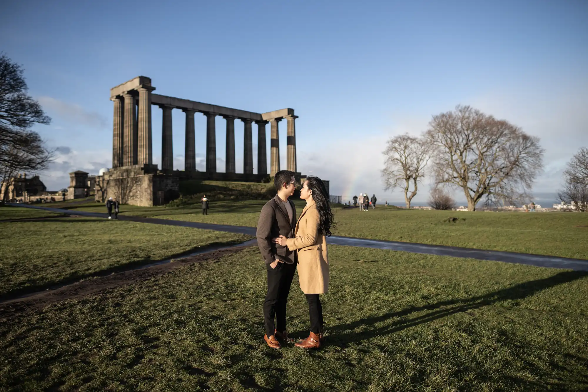 A couple stands facing each other, holding hands in an open grassy area. In the background, there is a large ancient structure with tall columns and a clear sky.