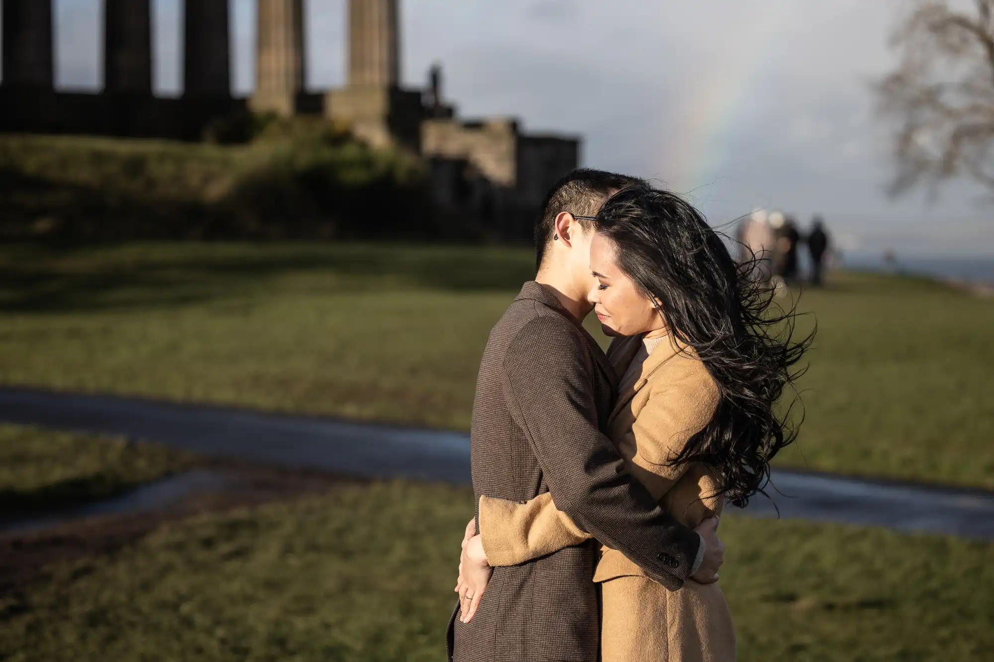 A couple embraces outdoors on a grassy area near historical ruins. A faint rainbow is visible in the background under a partly cloudy sky.