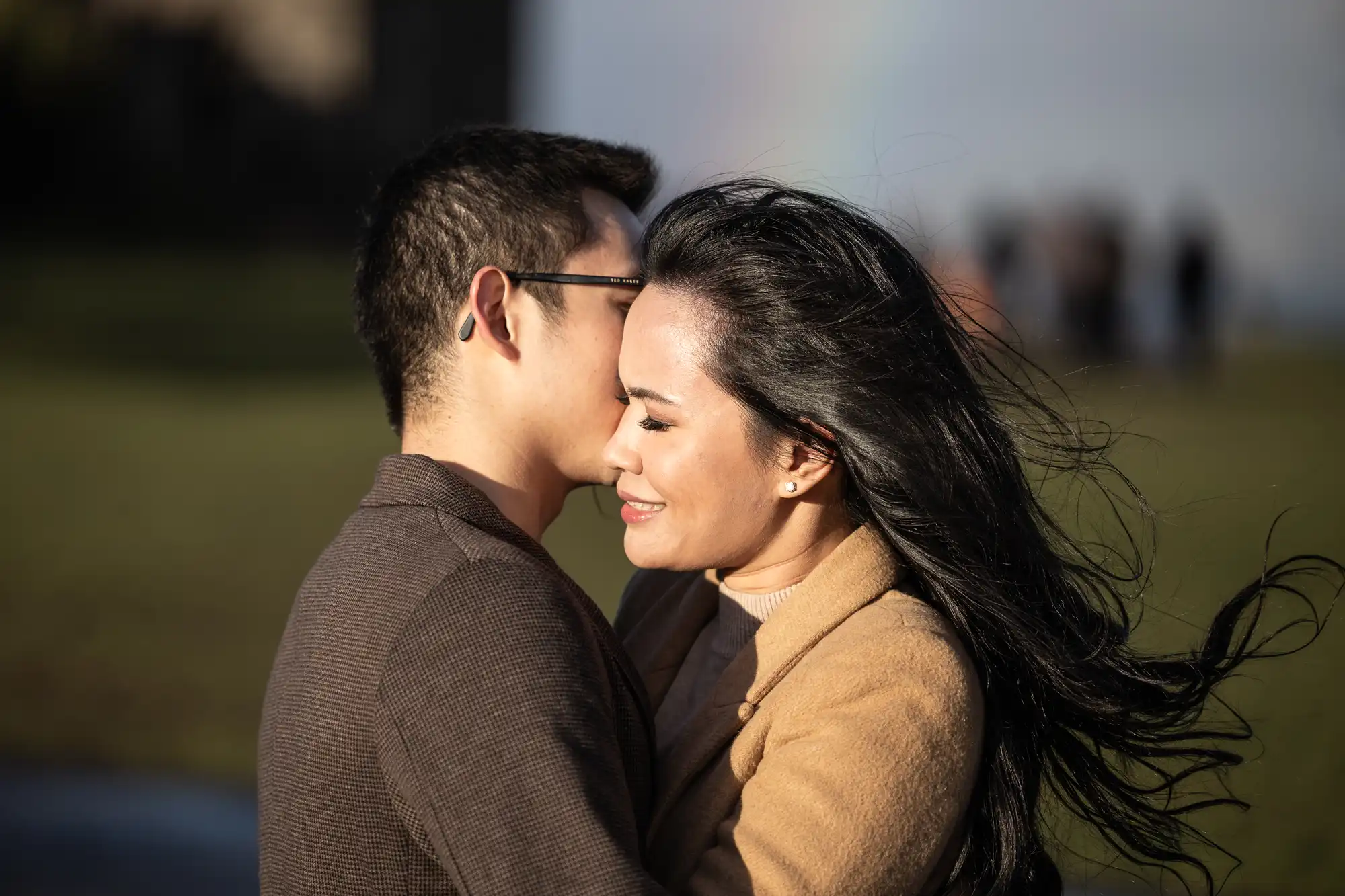 A couple embraces outdoors on a sunny day with a faint rainbow in the background. The woman faces the camera, smiling with eyes closed, while the man faces her, partially obscured.