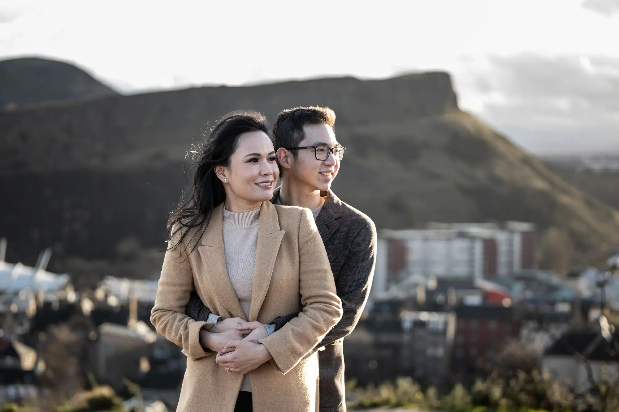 A woman and a man stand outdoors with mountainous terrain and buildings in the background. The woman is in front, both face to the right, and the man holds her from behind.