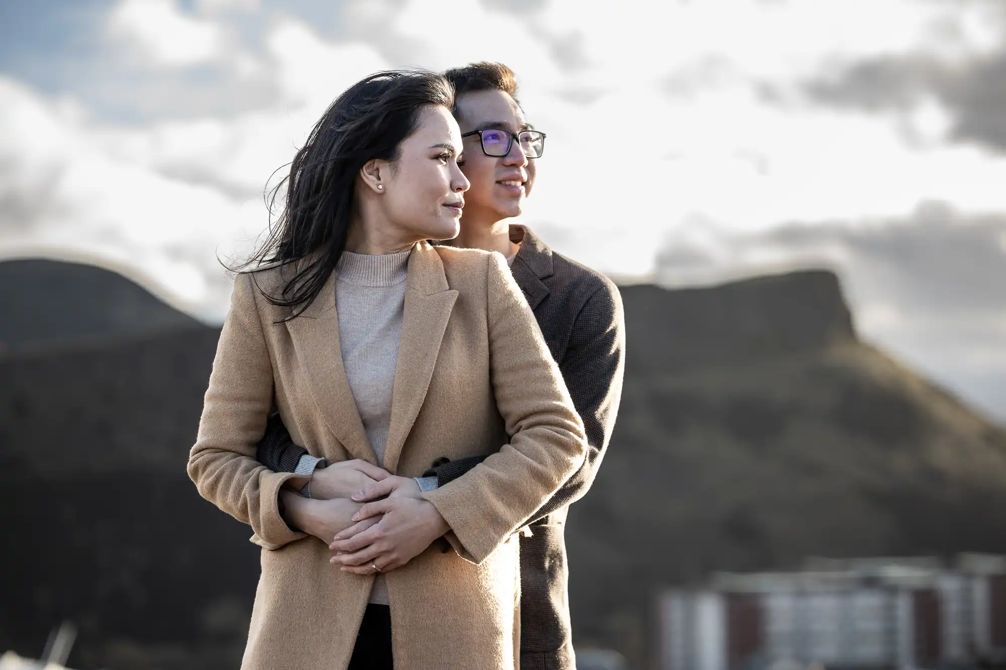 A couple stands outdoors with mountains in the background. The woman is in front, wearing a tan coat, while the man behind her, in glasses and a dark coat, embraces her and looks into the distance.